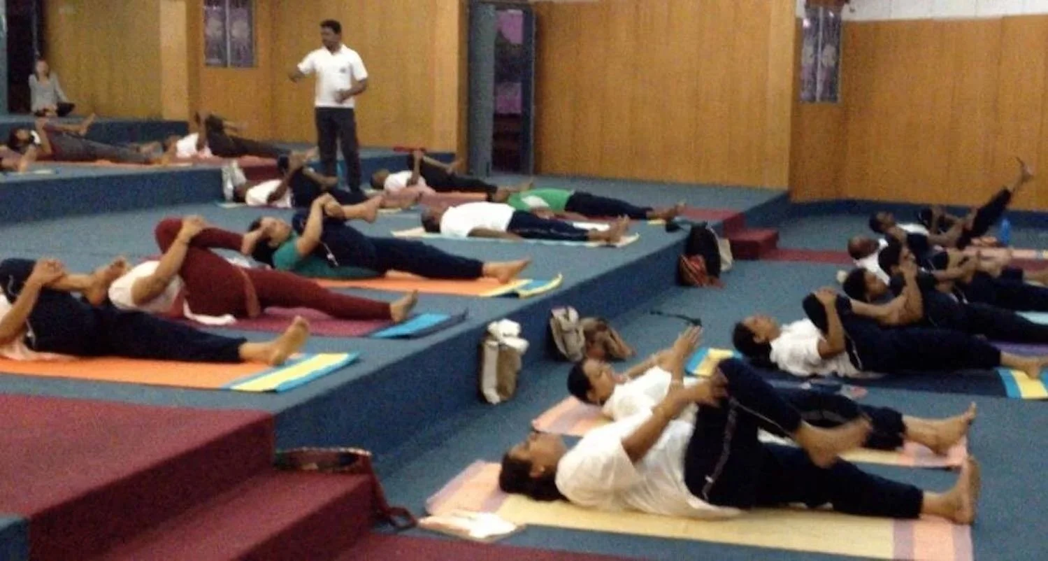 Group of people practicing yoga in a studio, lying on mats in a side stretch pose, instructor standing at the front.