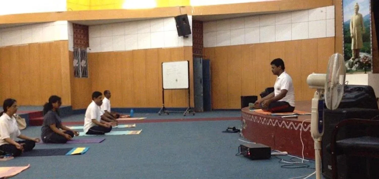 People sitting cross-legged on mats in a meditation classroom, with an instructor sitting in front of them on a raised platform.