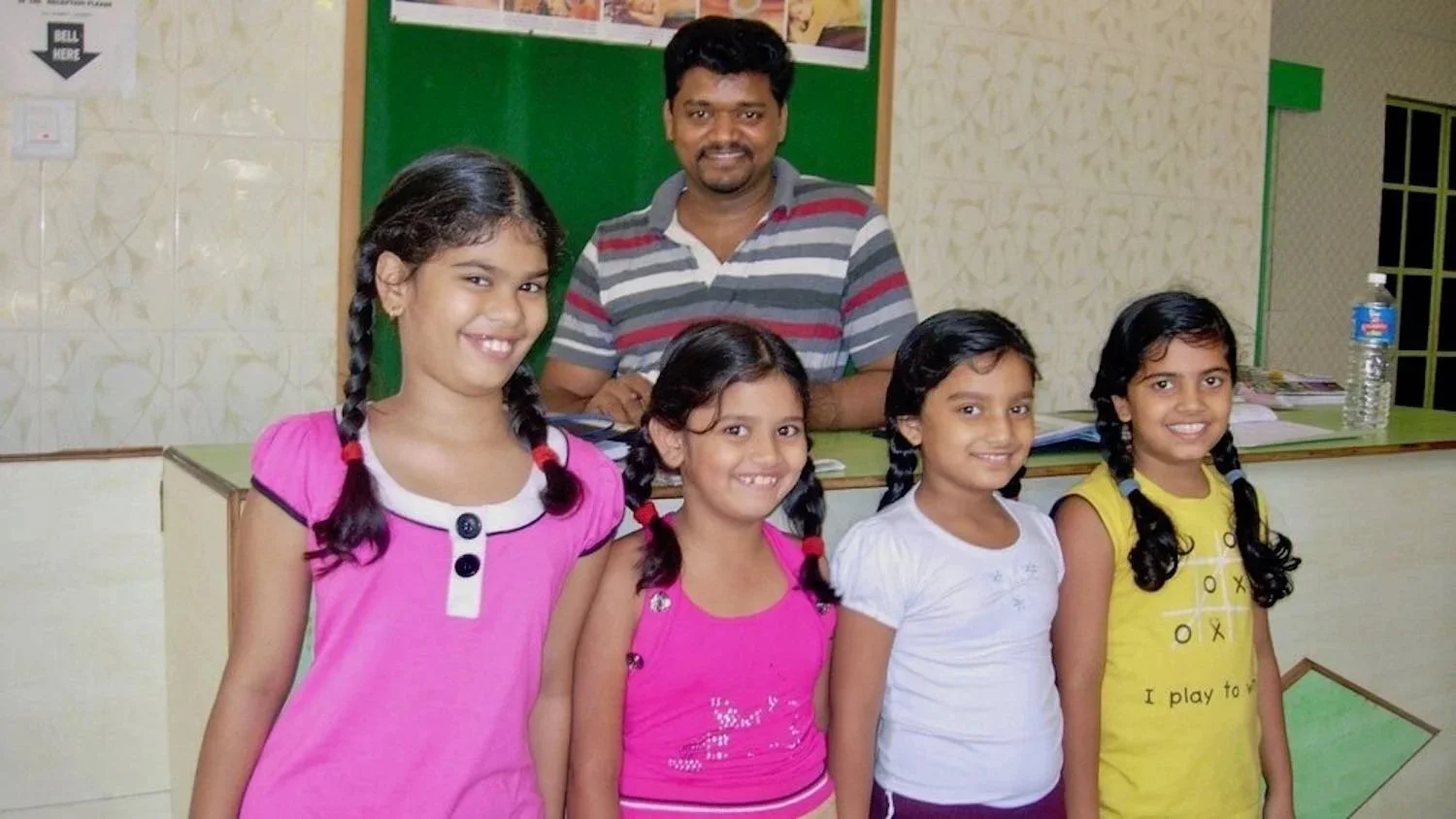 A man standing behind four young girls in a classroom or school setting, all smiling at the camera.
