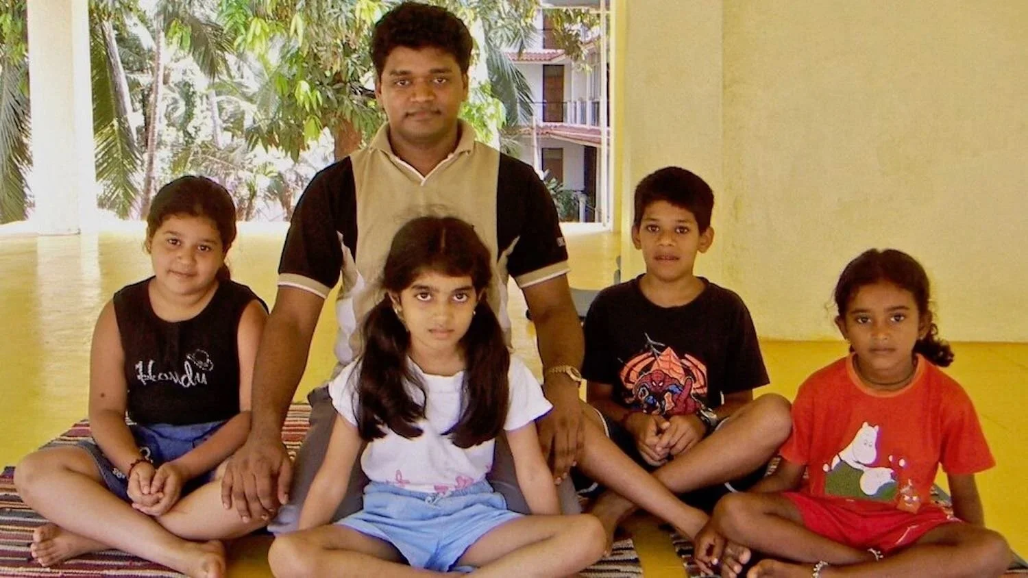 A man with five children sitting on a striped rug inside a yellow room, with trees visible through the window in the background.