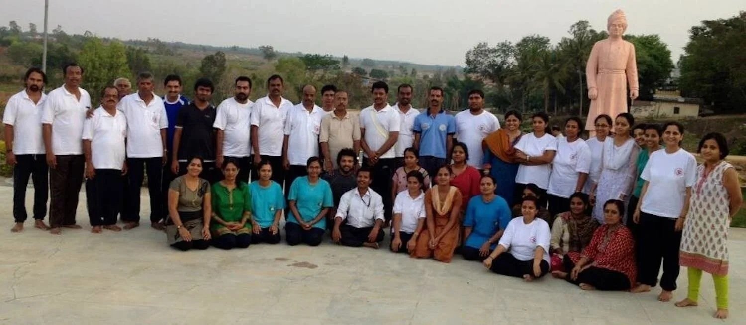 Group of people outdoors, standing and sitting in front of a large statue of a man, with trees and hills in the background.