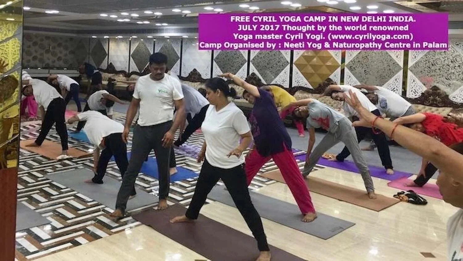 Group of people participating in a yoga class, performing side stretches on yoga mats in a decorated indoor space, with a signboard for a free yoga camp in New Delhi, India.