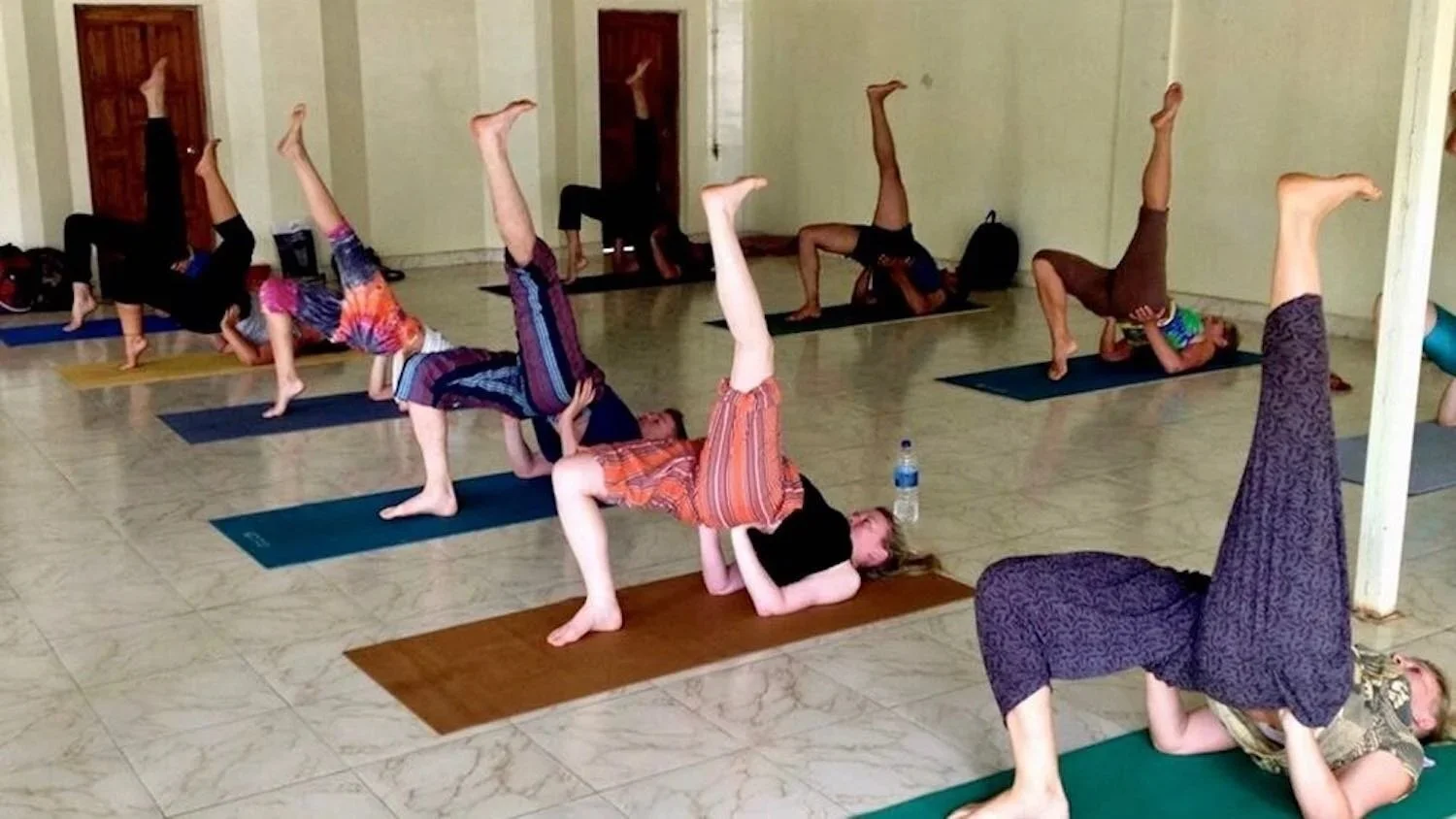A group of people engaging in a yoga class inside a room, performing a pose where they balance on their shoulders with one leg raised vertically, on yoga mats.