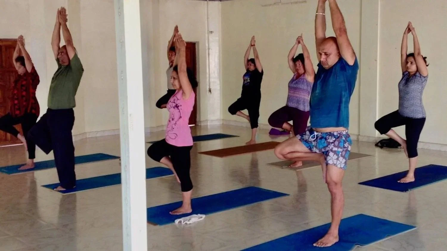 People participating in a yoga class, standing on yoga mats in a cross-legged pose with hands in prayer position overhead, inside a room with light-colored walls.