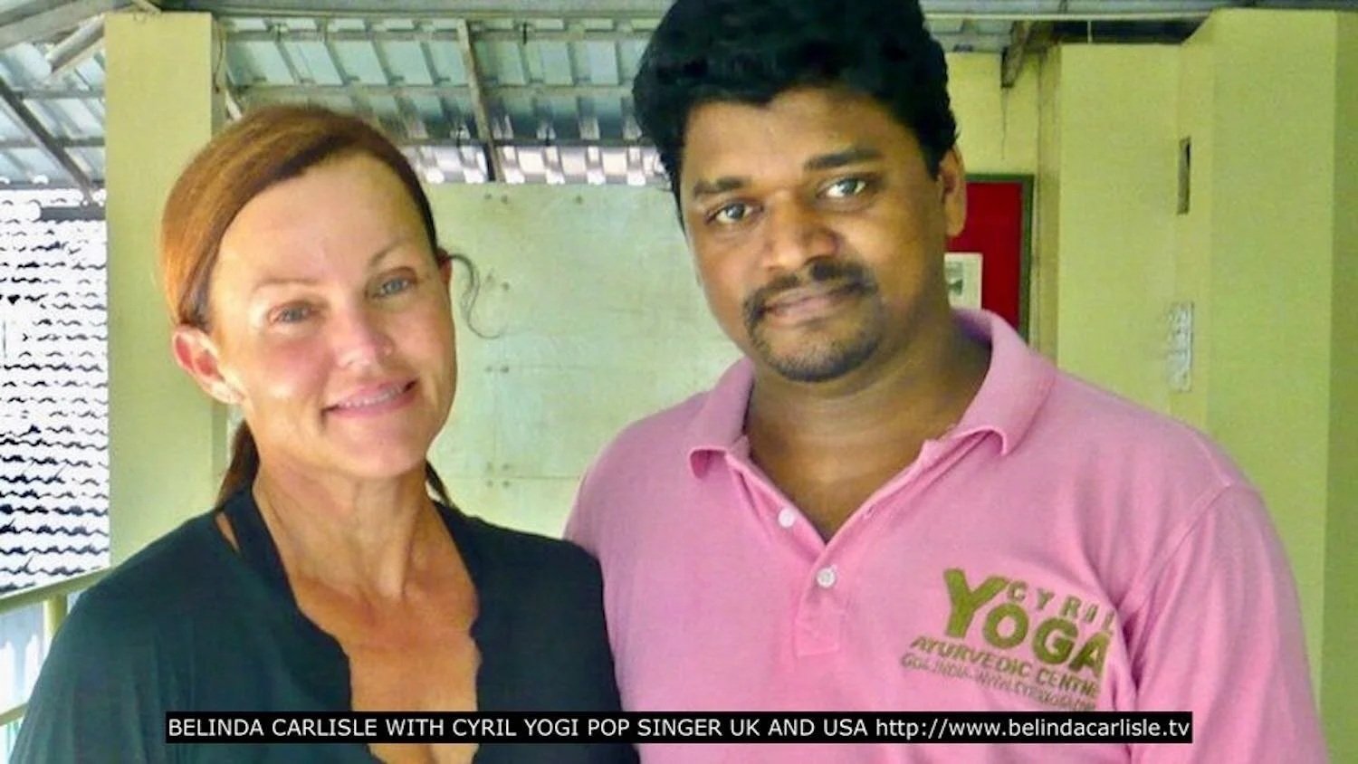 A woman with red hair and a man with black hair standing together indoors. The woman is wearing a black top, and the man is wearing a pink polo shirt with a logo. The background shows a yellow wall and a green partition.