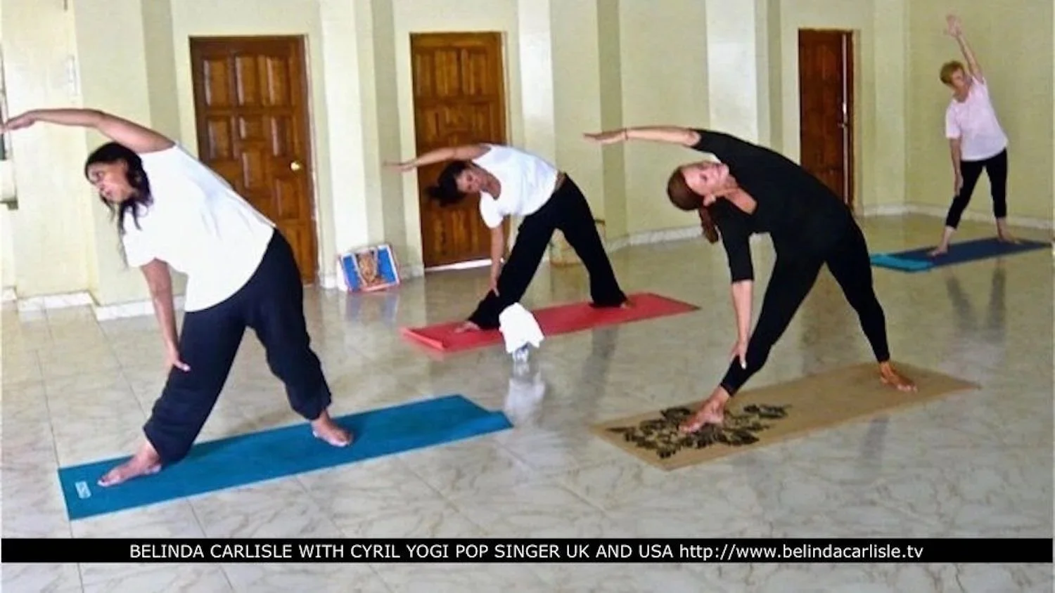 Four people practicing yoga in a room, all in side stretches with one arm over their head and the other touching their foot, on yoga mats of different colors.