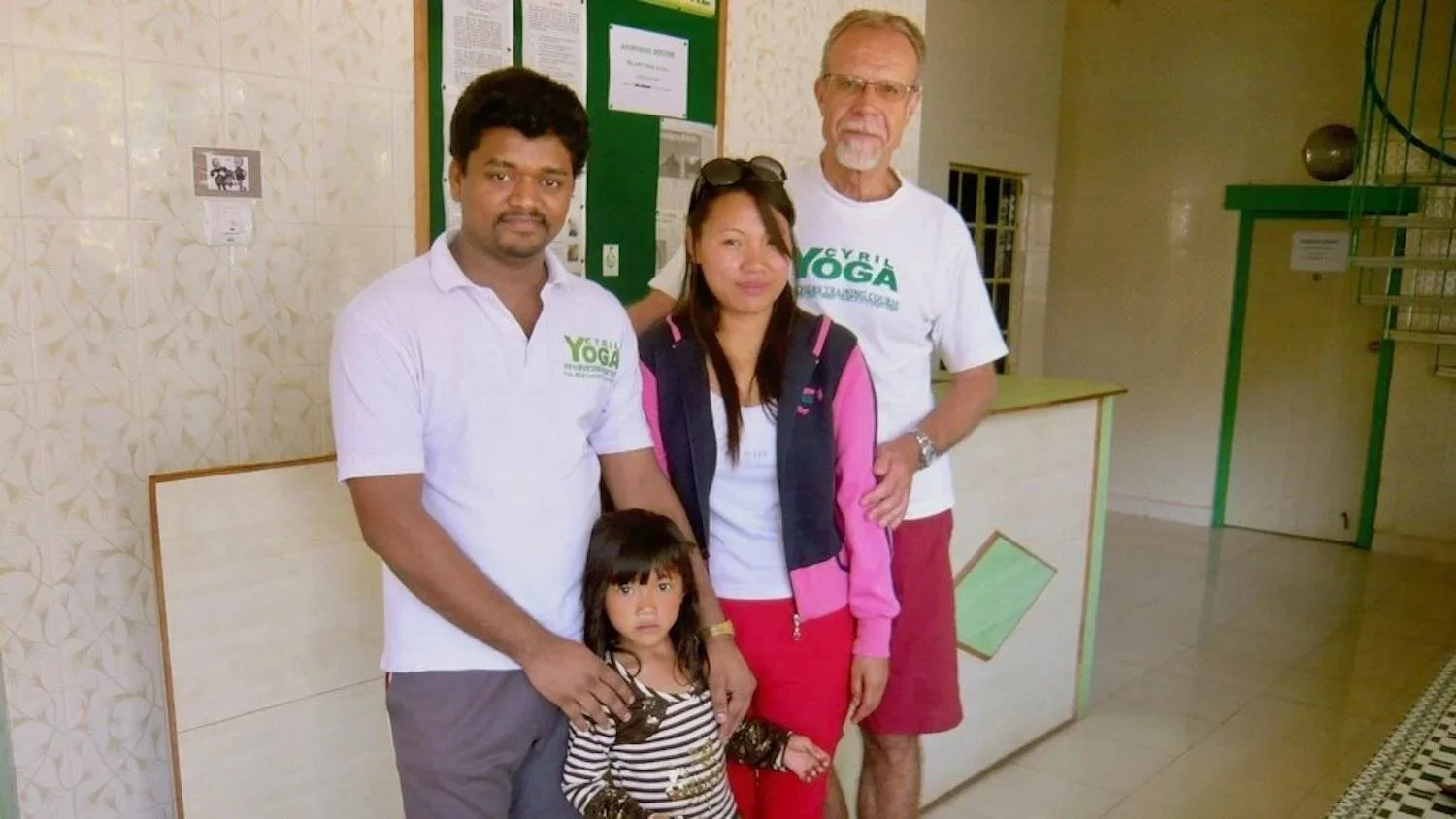 A group of four adults and one child posing inside a building, with two men wearing white T-shirts with a green YMCA logo, a woman in a pink and black jacket, and a young girl in a striped shirt.