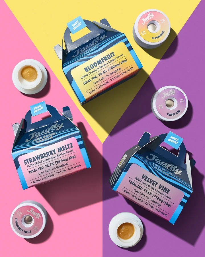 Three colorful boxes of cannabis edibles, labeled 'Strawberry Meltz,' 'Bloomfruit,' and 'Velvet Vine,' are placed on a vibrant background with assorted small containers of cannabis concentrates or extracts nearby.