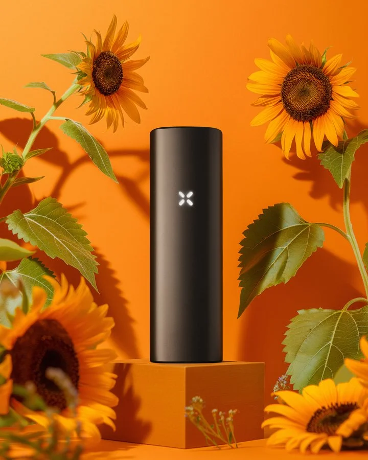 A black cylindrical device with a glowing white logo stands on an orange pedestal, surrounded by sunflowers with large green leaves, set against an orange background.