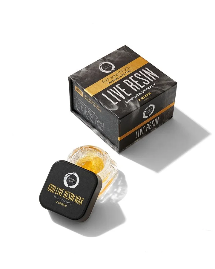 A black box labeled 'CBD Live Resin Wax' with a small container of yellow wax inside, placed in front of a larger black box labeled 'Live Resin' containing cannabis extract, on a white background.