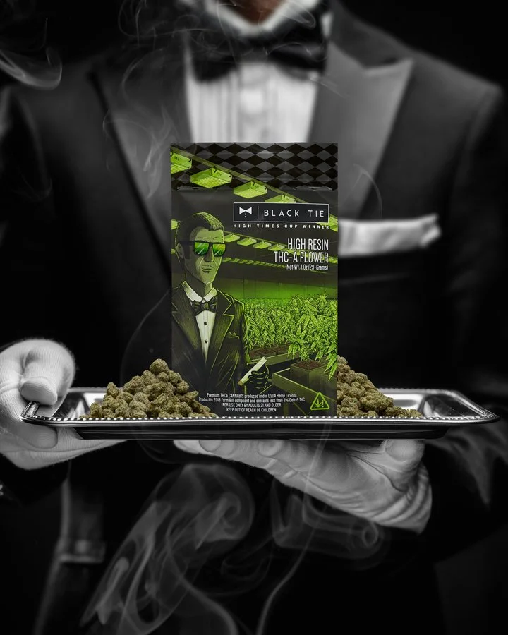 A man in a tuxedo holding a tray with cannabis buds and a box of high resin THC-A flower packaging, with a black-and-white background. The packaging features a cartoon man in sunglasses, a tuxedo, and a cityscape with marijuana plants.