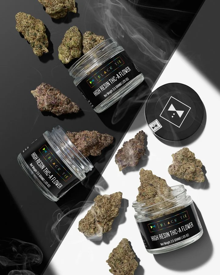 Three open containers of Black Tie high resin THC-A flowers cannabis buds with several buds scattered on a black and white surface, some with visible vapor.
