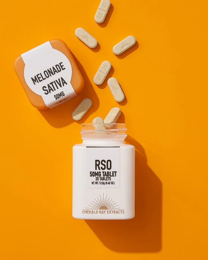 Open white bottle of RSO 50mg THC tablets with some tablets spilling out onto orange surface, alongside a larger container labeled Melonade Sativa 50MG, both on bright orange background.