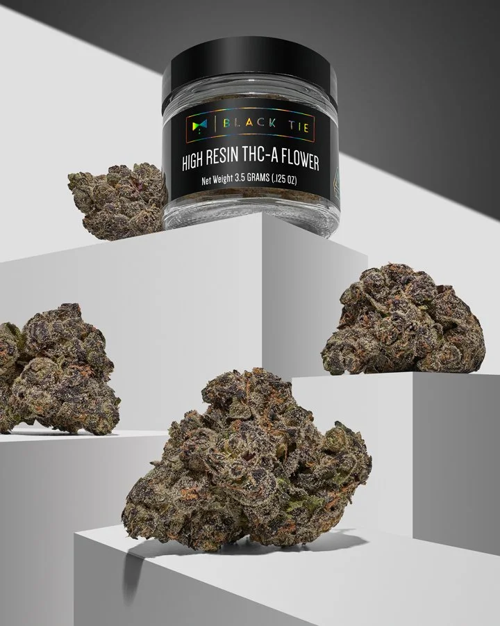 A jar labeled 'Black Tie High Resin THC-A Flower' surrounded by several large cannabis buds on white and gray geometric platforms.