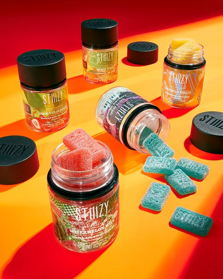 Several jars and containers of colorful gummy candies on an orange surface with a red background. Some jars are open and candies are spilling out.