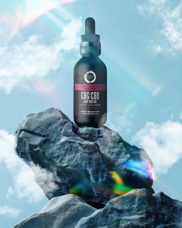 A bottle of CBD and THC hemp tincture with metta hemp label, placed on top of a large rock formation against a cloudy sky background with rainbow light reflections.