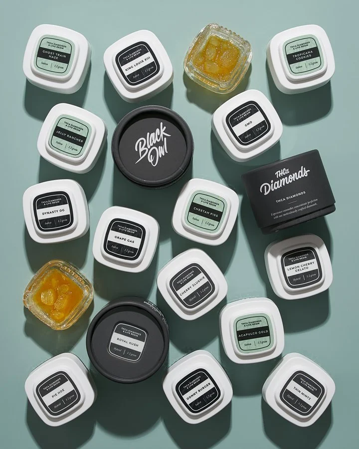 Various cannabis concentrate containers with different strain labels, two with yellow concentrates, a black grinder labeled 'Black Out', and a black container labeled 'THCa Diamonds', arranged on a teal surface.