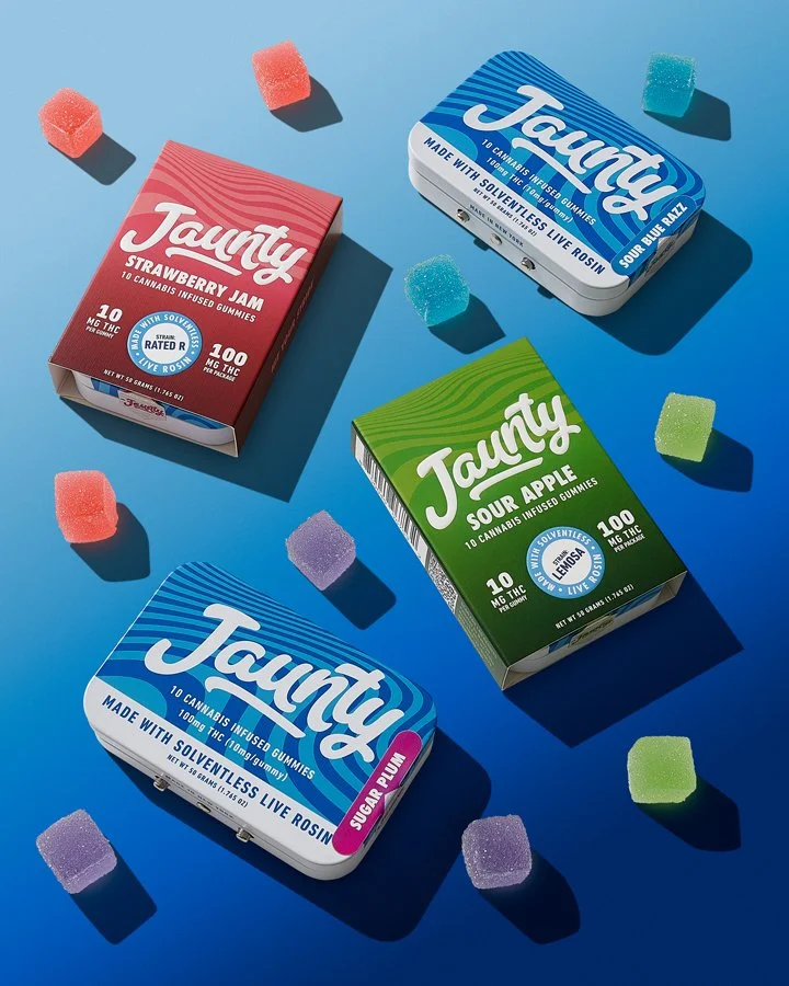 Three boxes of Tauntly cannabis-infused gummies in strawberry jam, sour apple, and sour blue raspberry flavors, with colorful gummy candies scattered around on a blue background.
