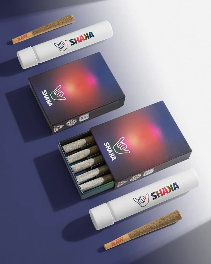 Two boxes of pre-rolled cannabis joints, two jars labeled 'SHAYA' with colorful logos, and two unlit raw cannabis joints, all arranged on a table.