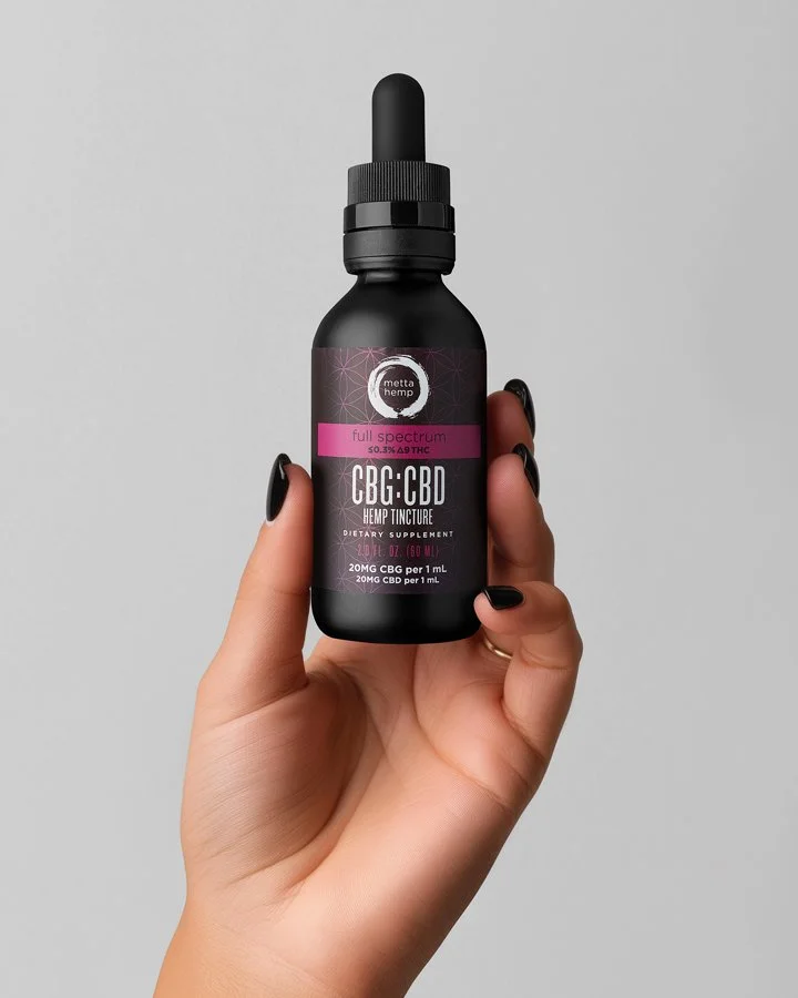 Hand holding a black dropper bottle of CBD hemp tincture labeled 'metta hemp' and 'full spectrum' with a gray background.