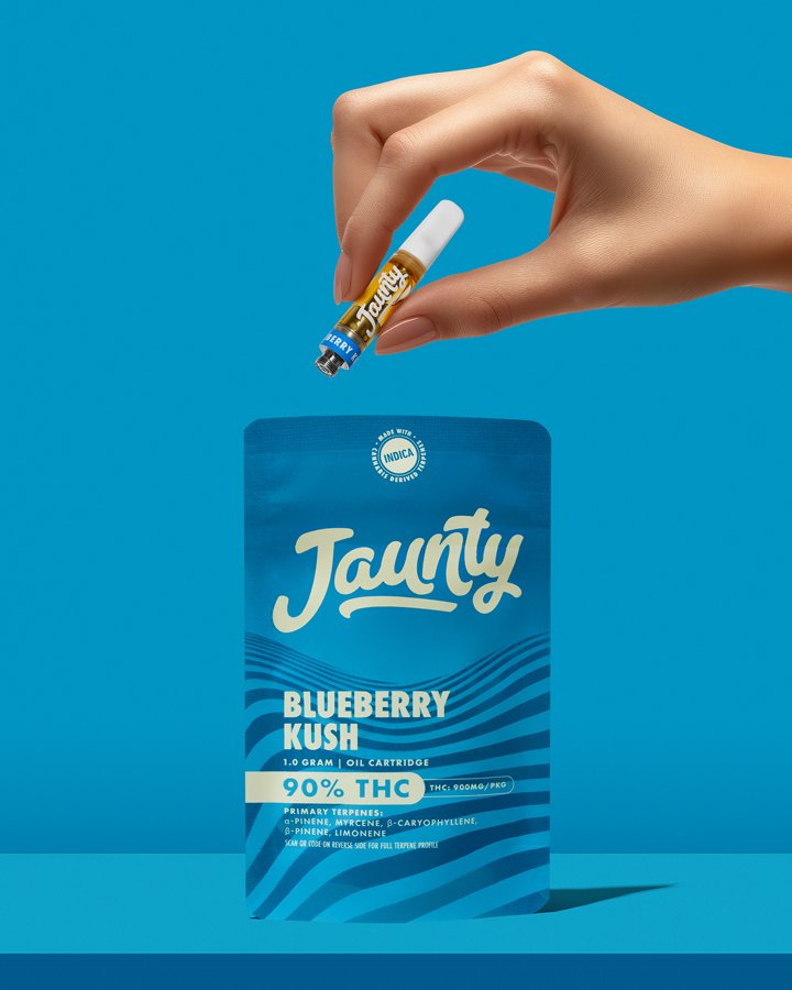 A hand holding a small vape cartridge labeled 'Taunt', with the tip positioned above a blue package labeled 'Blueberry Kush' containing cannabis oil, set against a blue background.