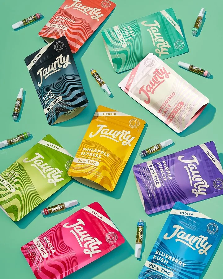 Various colorful packages of Jaunt cannabis products and small vape cartridges scattered on a mint green surface.