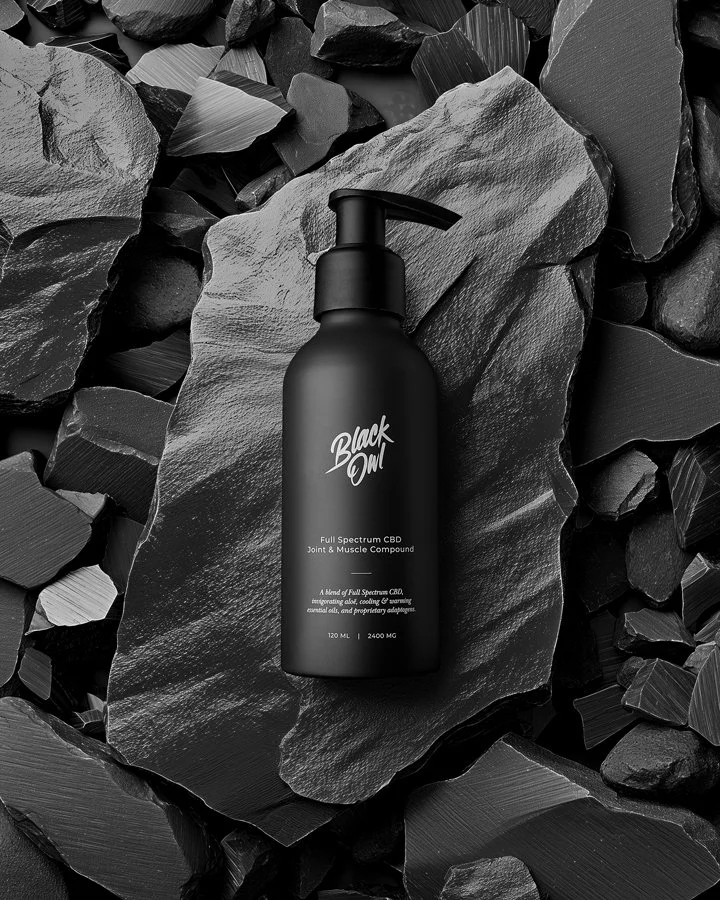 A black bottle with a pump labeled 'Black Owl' sits on a piece of slate surrounded by black rocks.