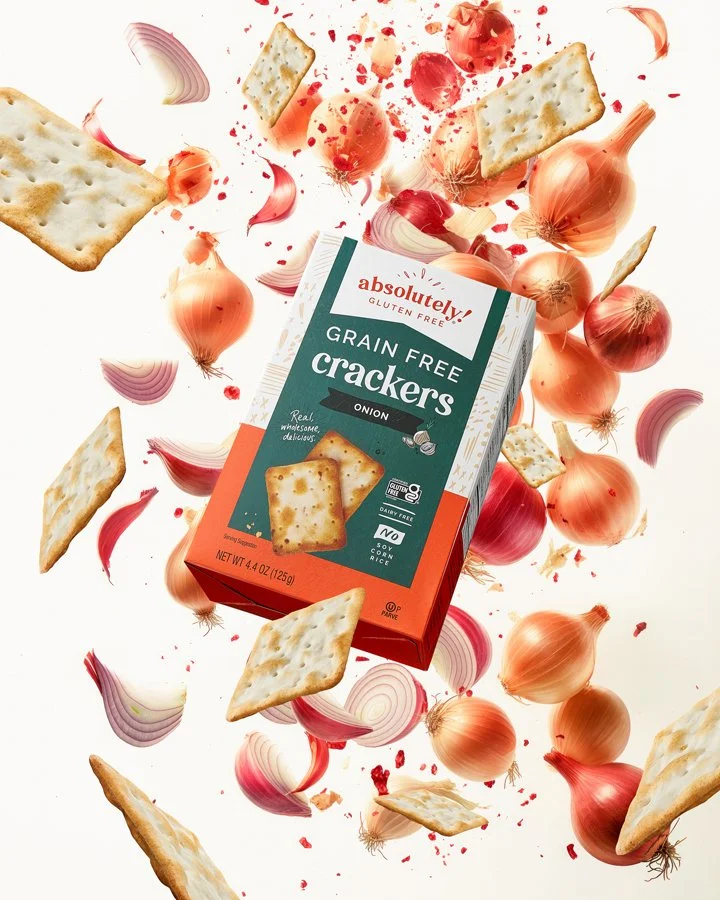 A box of Absolutely! gluten-free onion grain-free crackers surrounded by onion slices and fragments flying around.