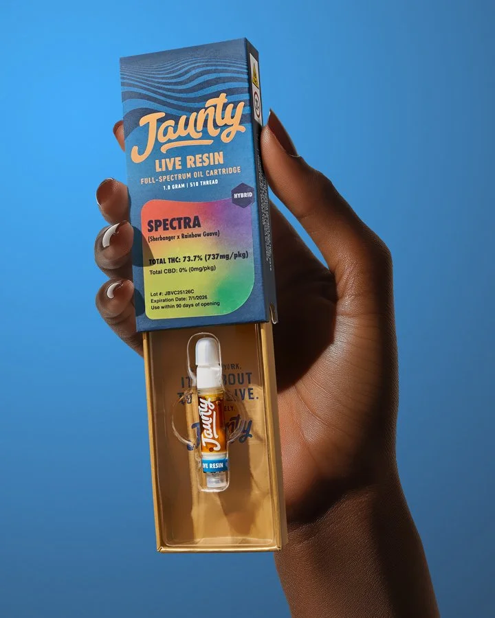 A hand holding a box of Taunty live resin in front of a blue background. The box is blue with orange and rainbow accents, highlighting the product's THC and CBD content.