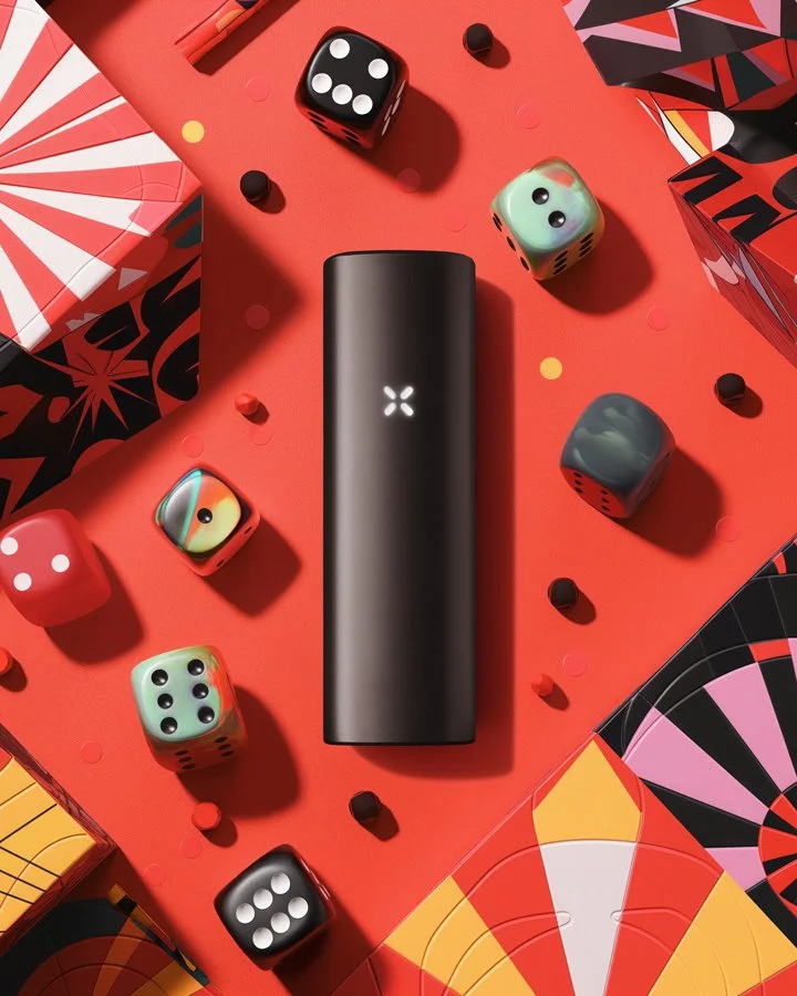 A black vape device is centered on a red background surrounded by colorful dice and patterned boxes, with scattered small round confetti.