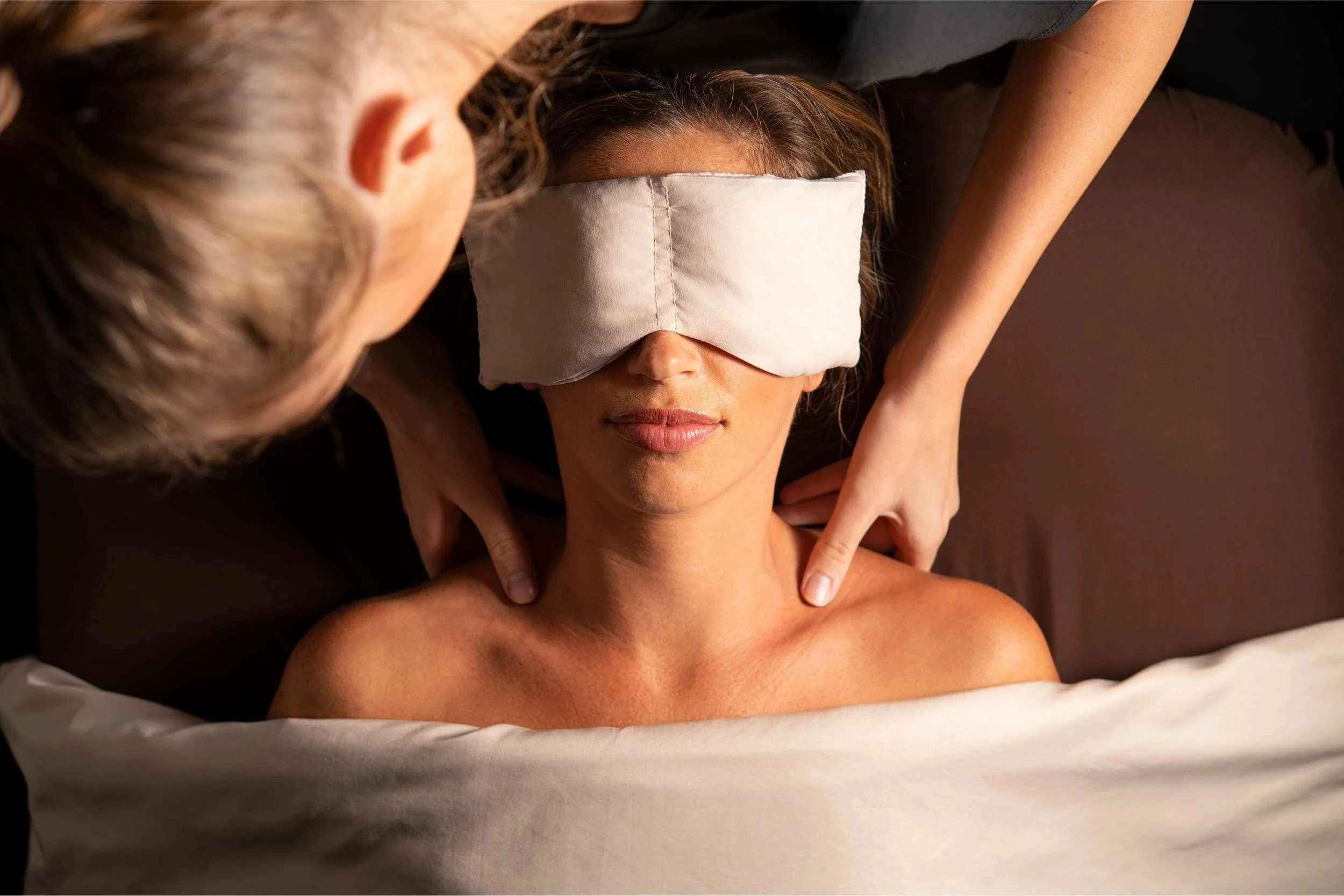A woman wearing an eye mask receives a massage for self care, the massage therapist massages her shoulders