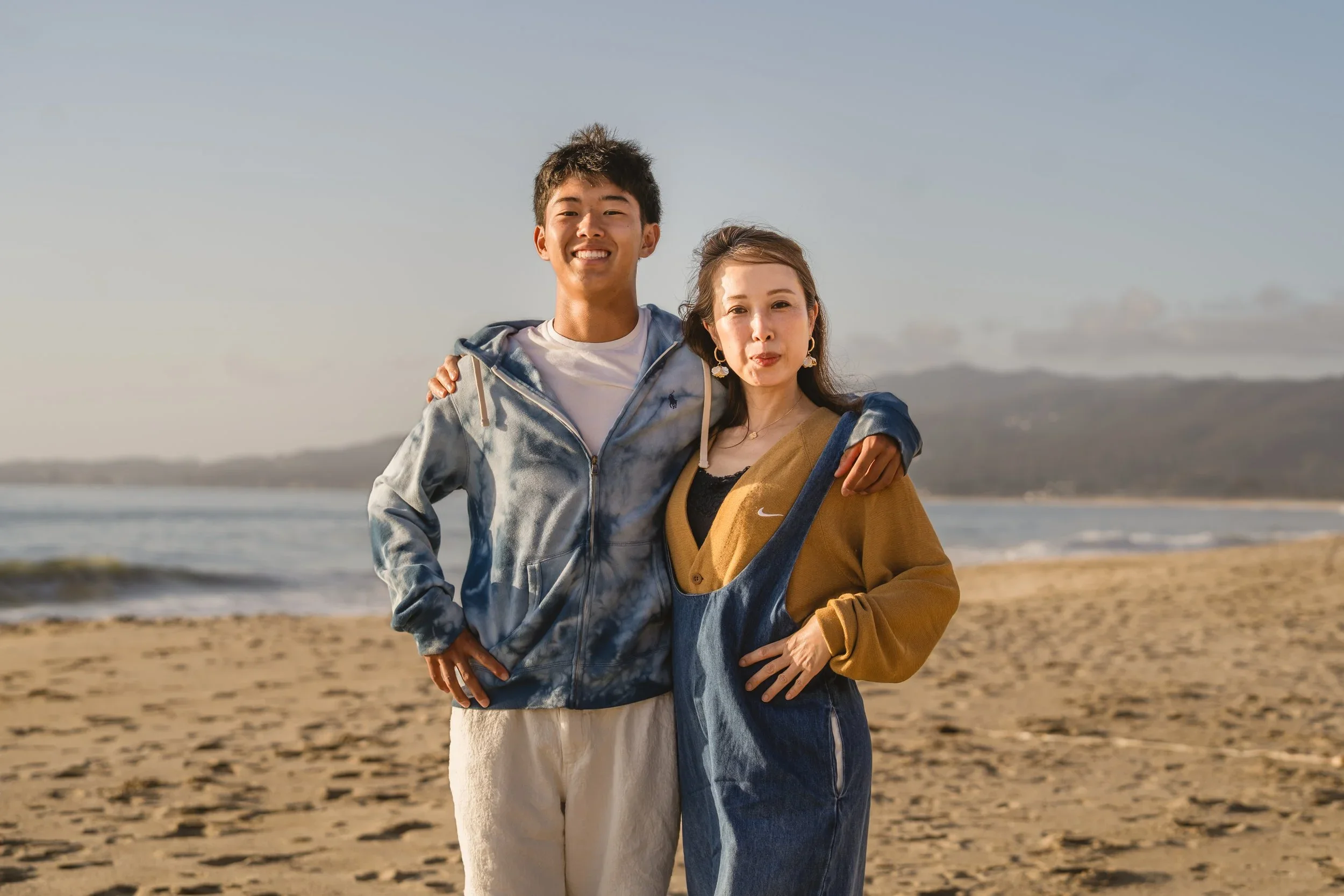 Mother and her son stand arm in arm on the sand at Poplar Beach, with waves and coastal hills behind them in late afternoon light