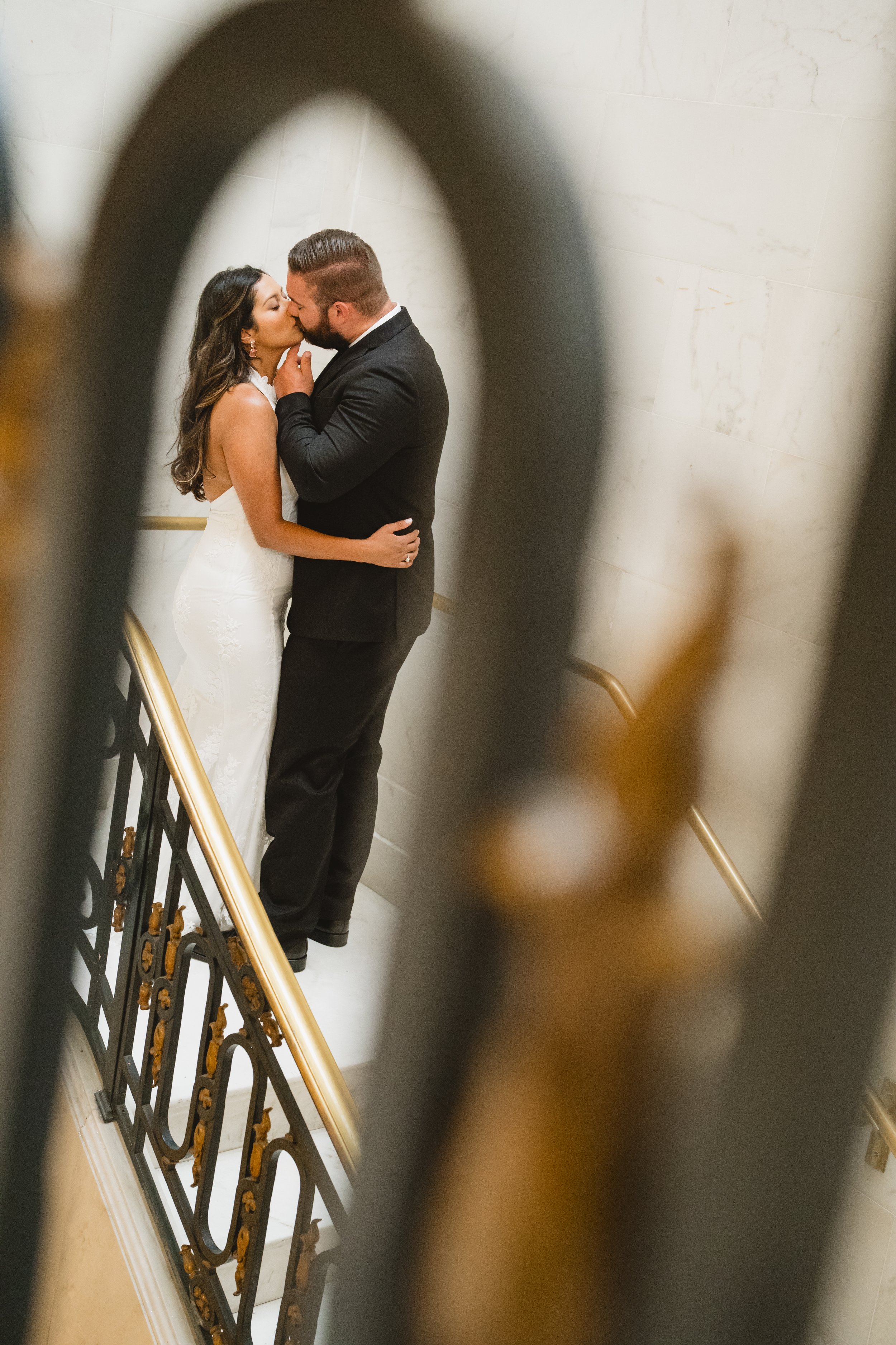 Couple kissing on a staircase, viewed through the decorative ironwork of a stair railing.