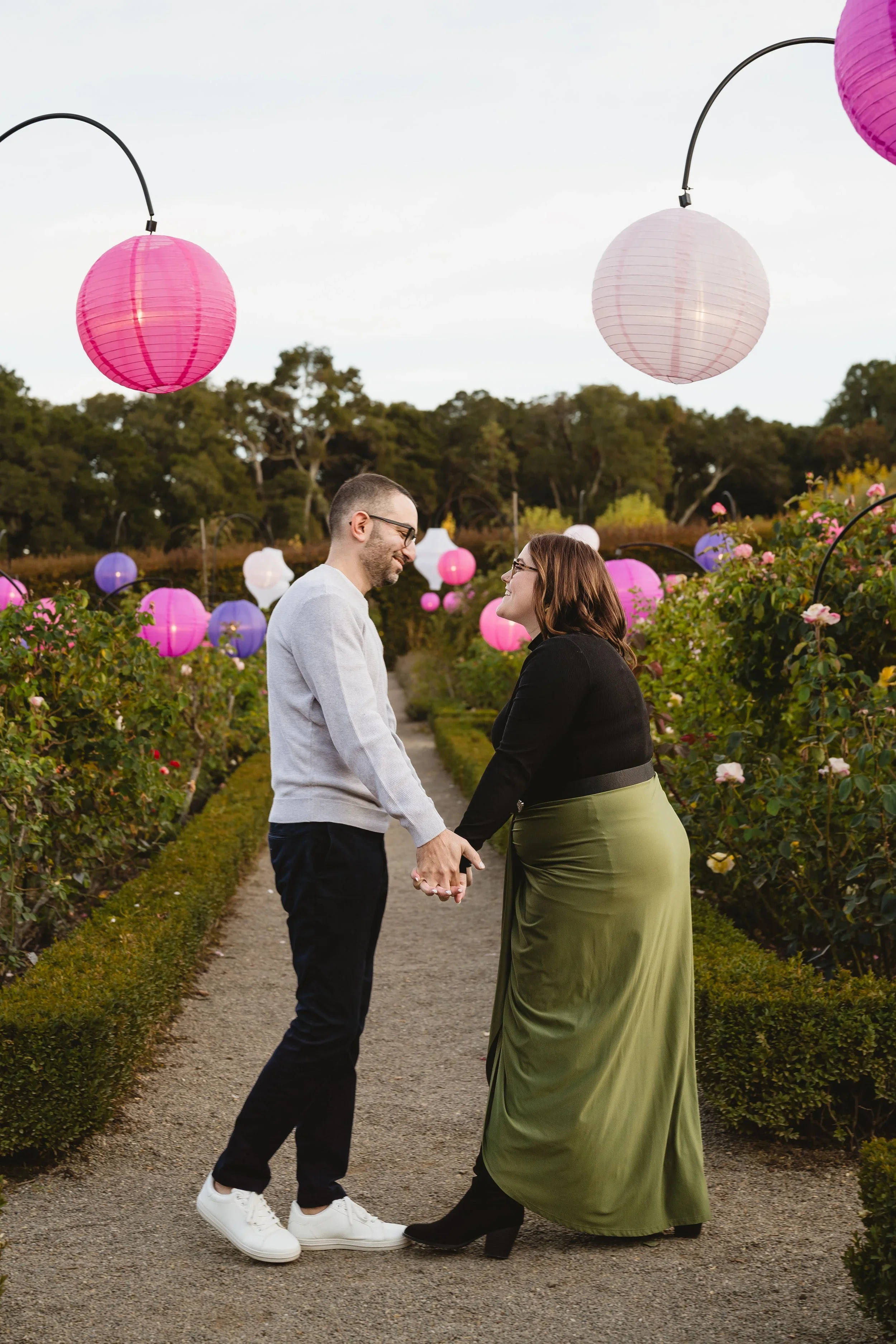 Couple facing each other and holding hands on a gravel rose garden path lined with hanging pink and purple paper lanterns at Filoli.