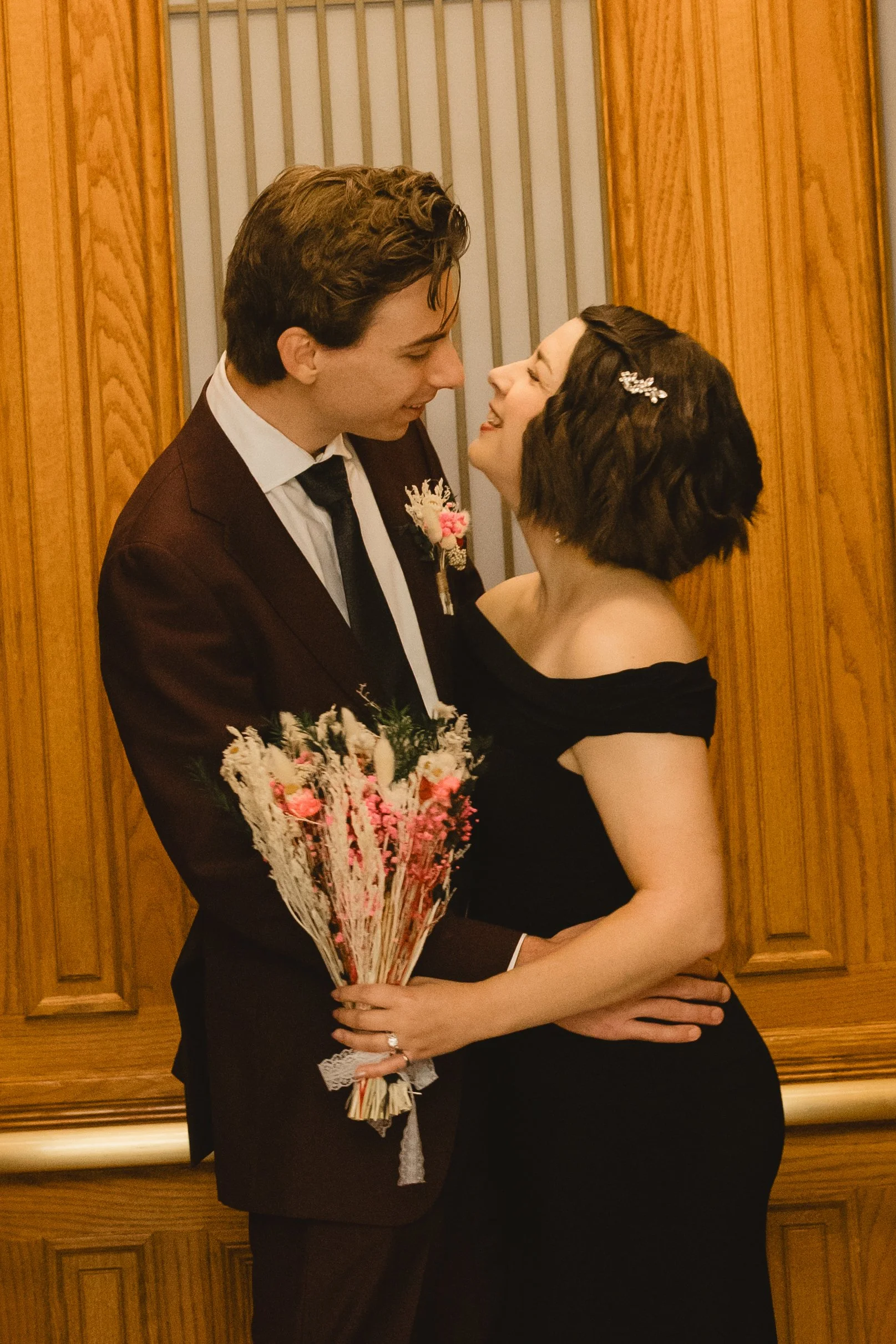 A couple dressed in formal attire embracing each other, the woman holding a bouquet of pink and white flowers, standing in front of wooden paneling.