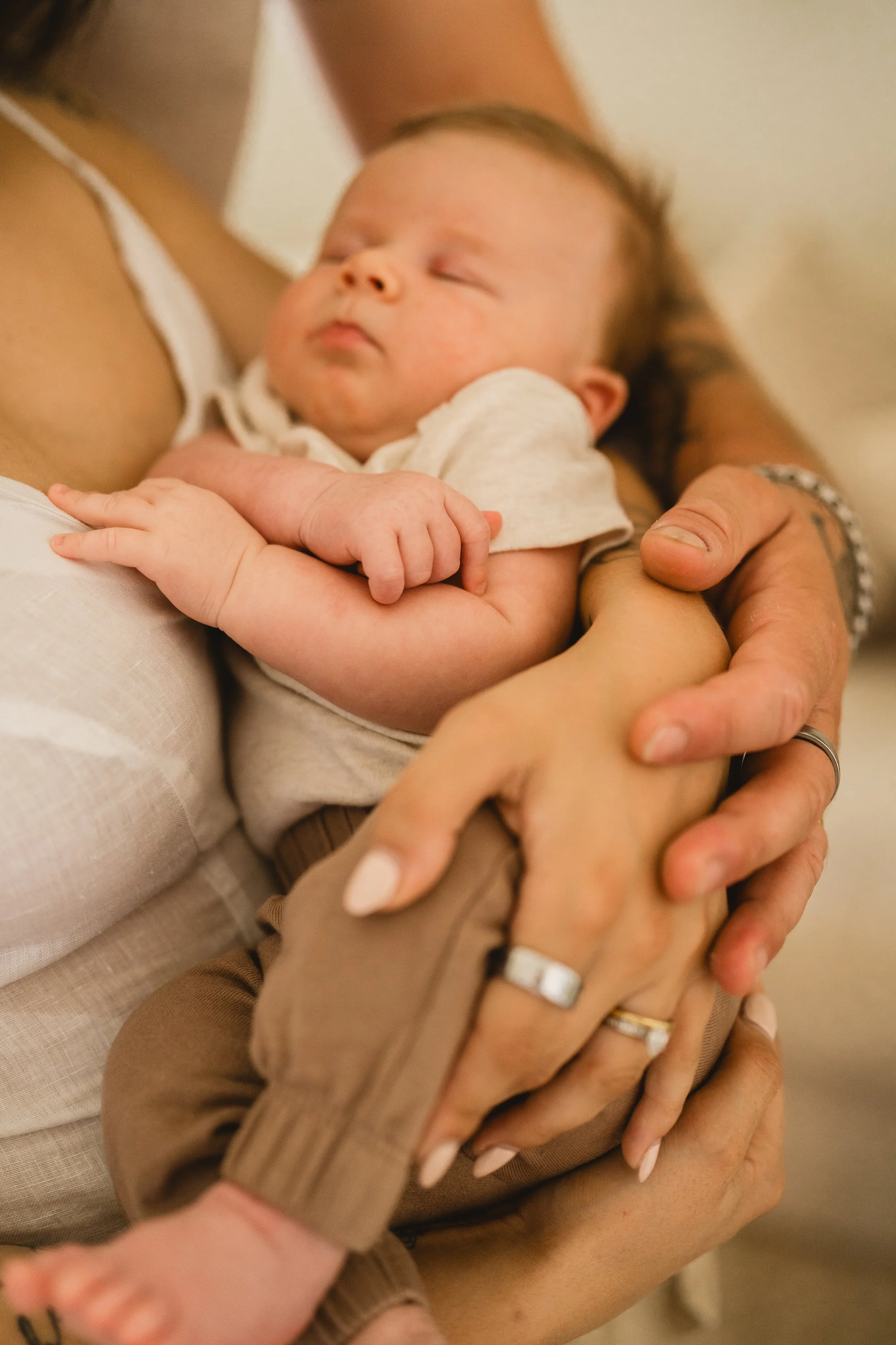 A newborn sleeps in a parent's arms while both parents' ringed hands rest stacked over the baby's legs in a close, cropped frame