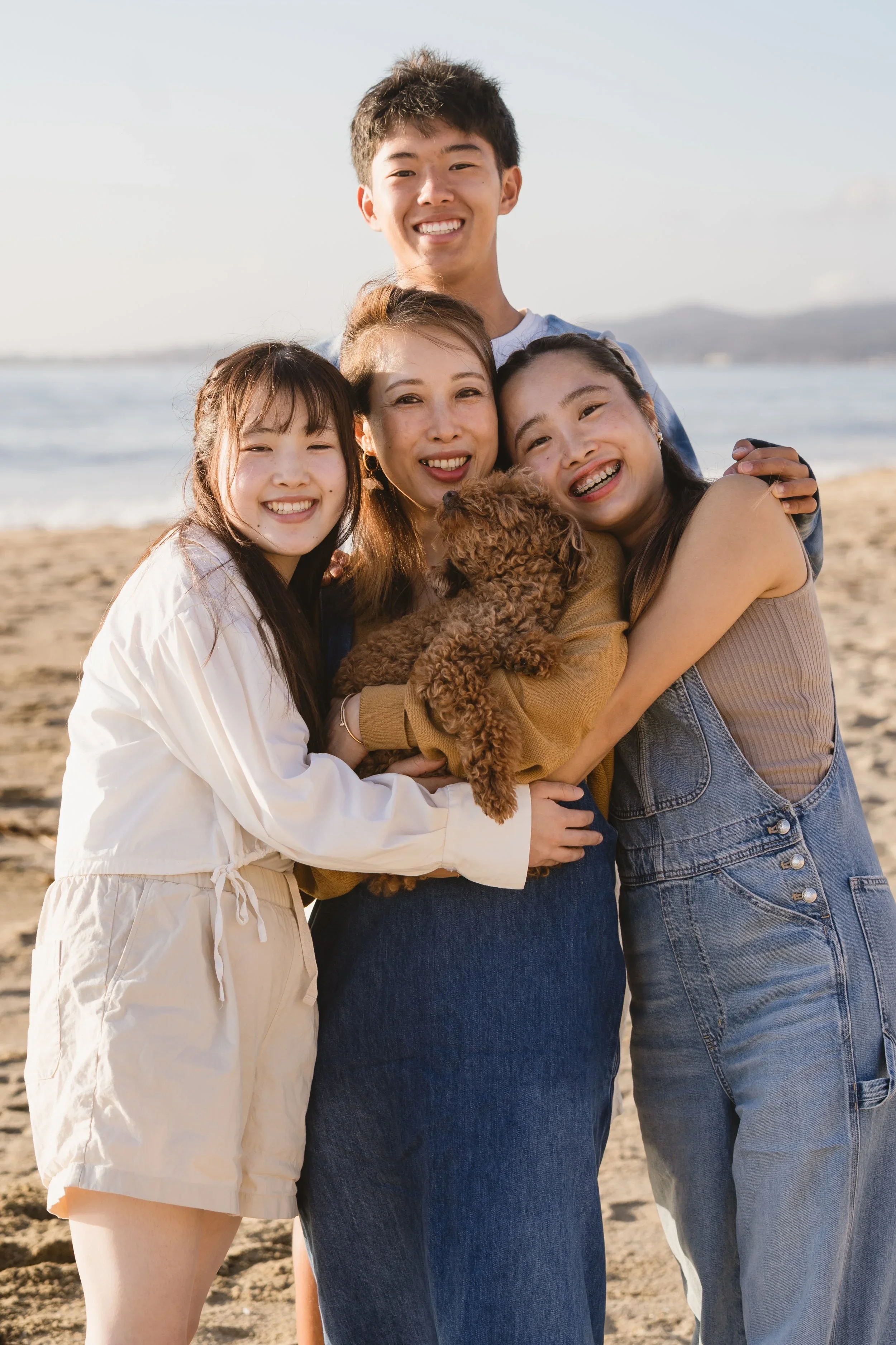 Mom holds the family poodle mix at the center while her son and two daughters lean in around her, ocean and open sand behind them