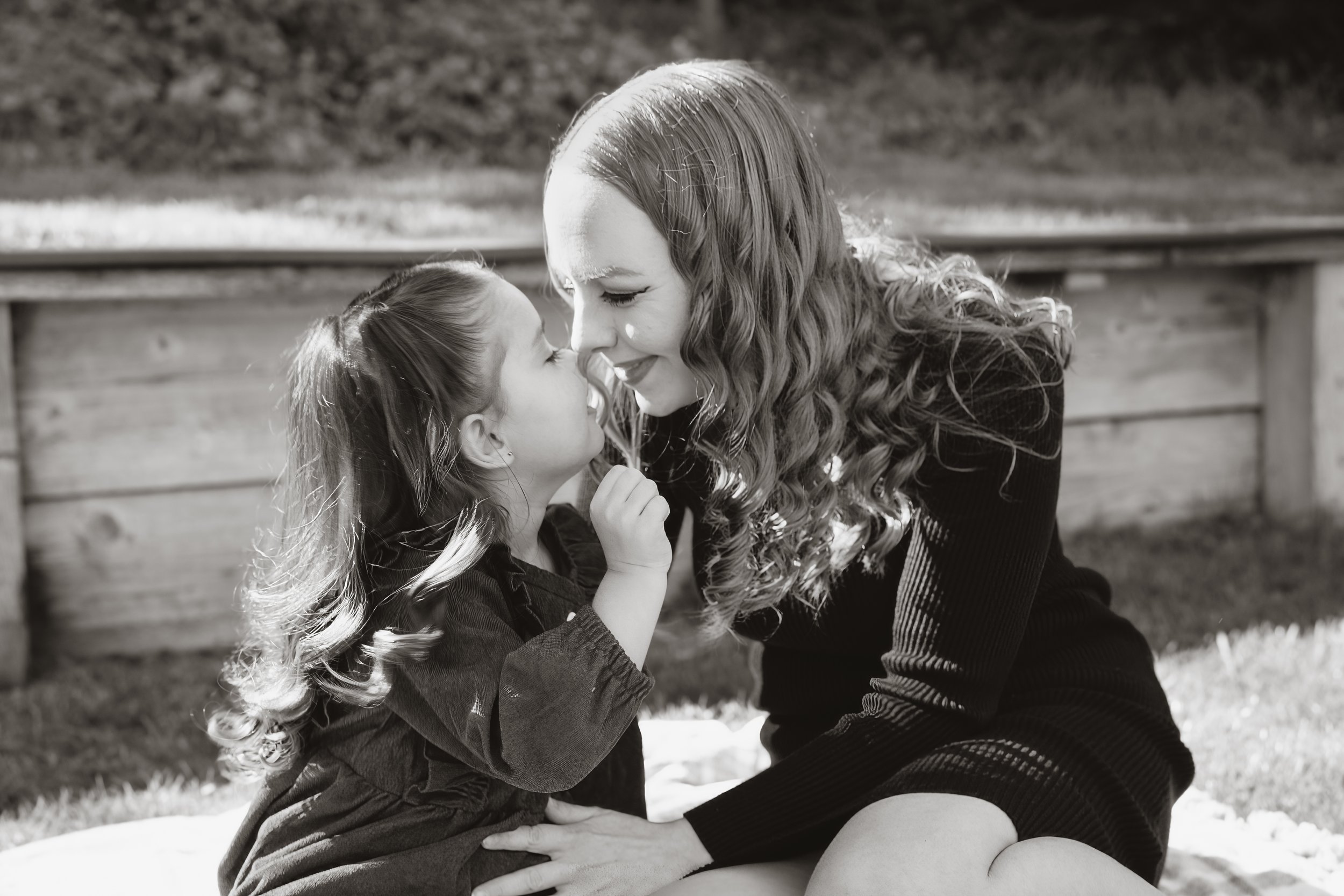 A woman and a girl with curly hair share a tender kiss outdoors, sitting on the grass with a wooden fence behind them.