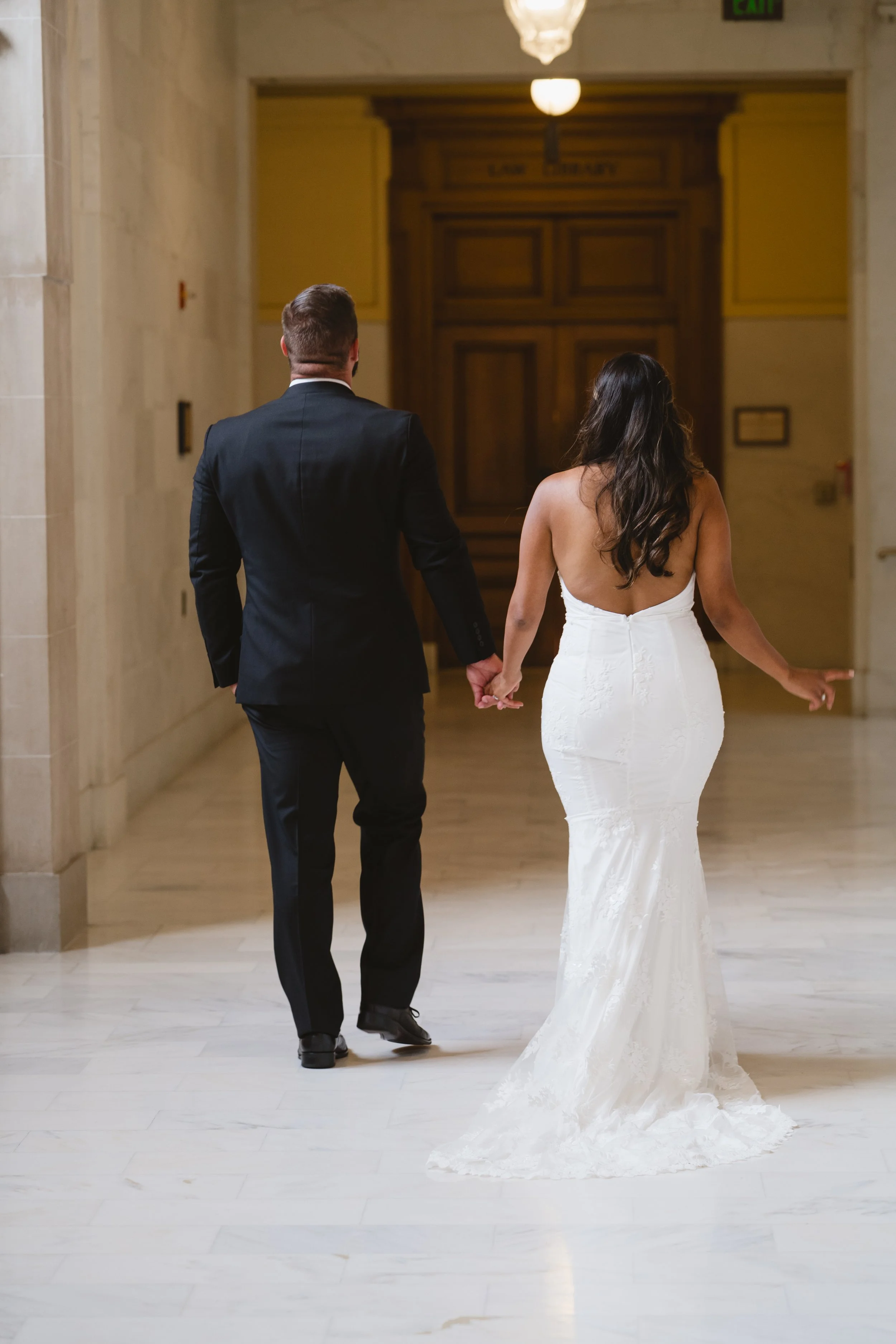 Back view of the couple walking hand in hand down a marble corridor toward a wood-paneled door.