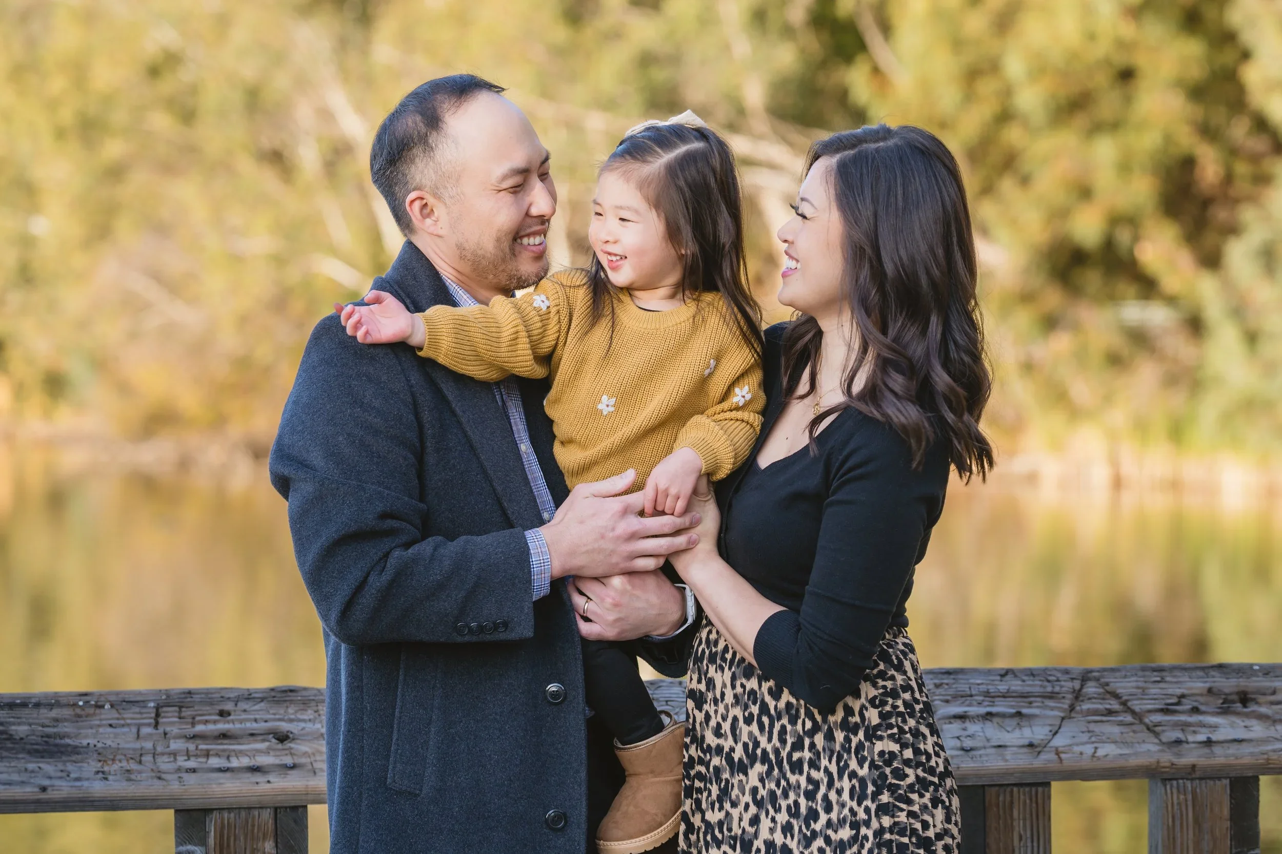 On the bridge at Vasona Lake, a father and mother laugh with their daughter as she reaches toward him, all looking at each other rather than the camera.