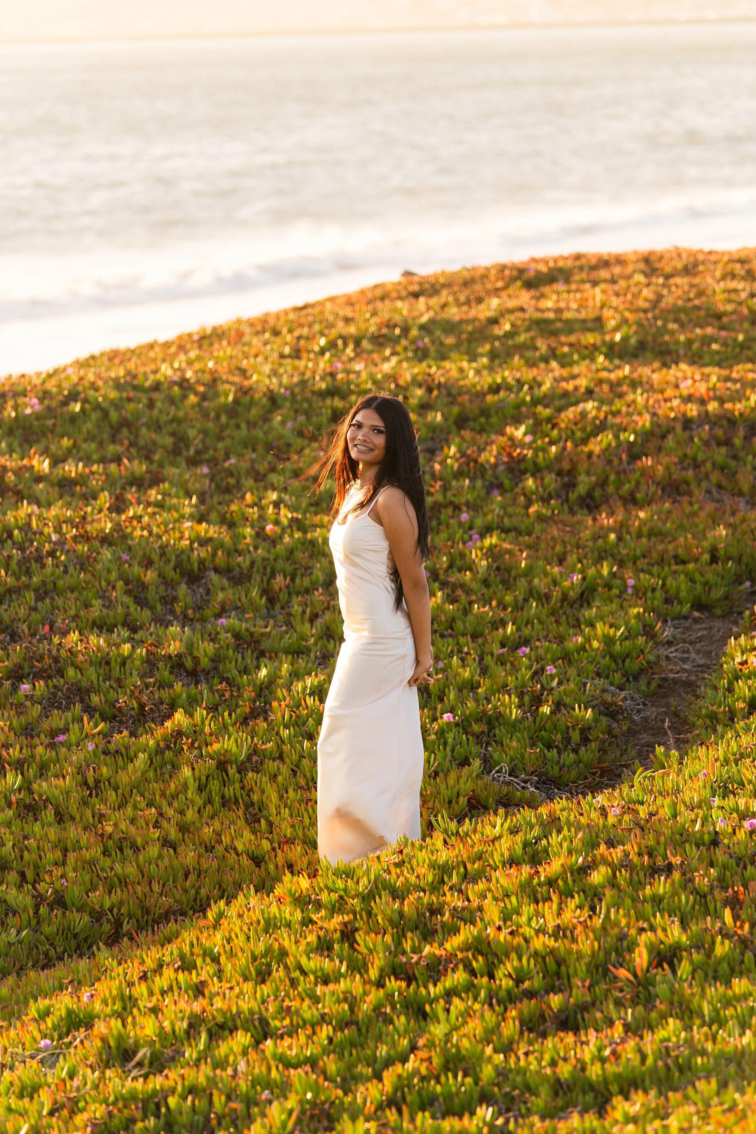 Senior in a cream dress standing on a green and orange ice plant bluff above the ocean in Half Moon Bay