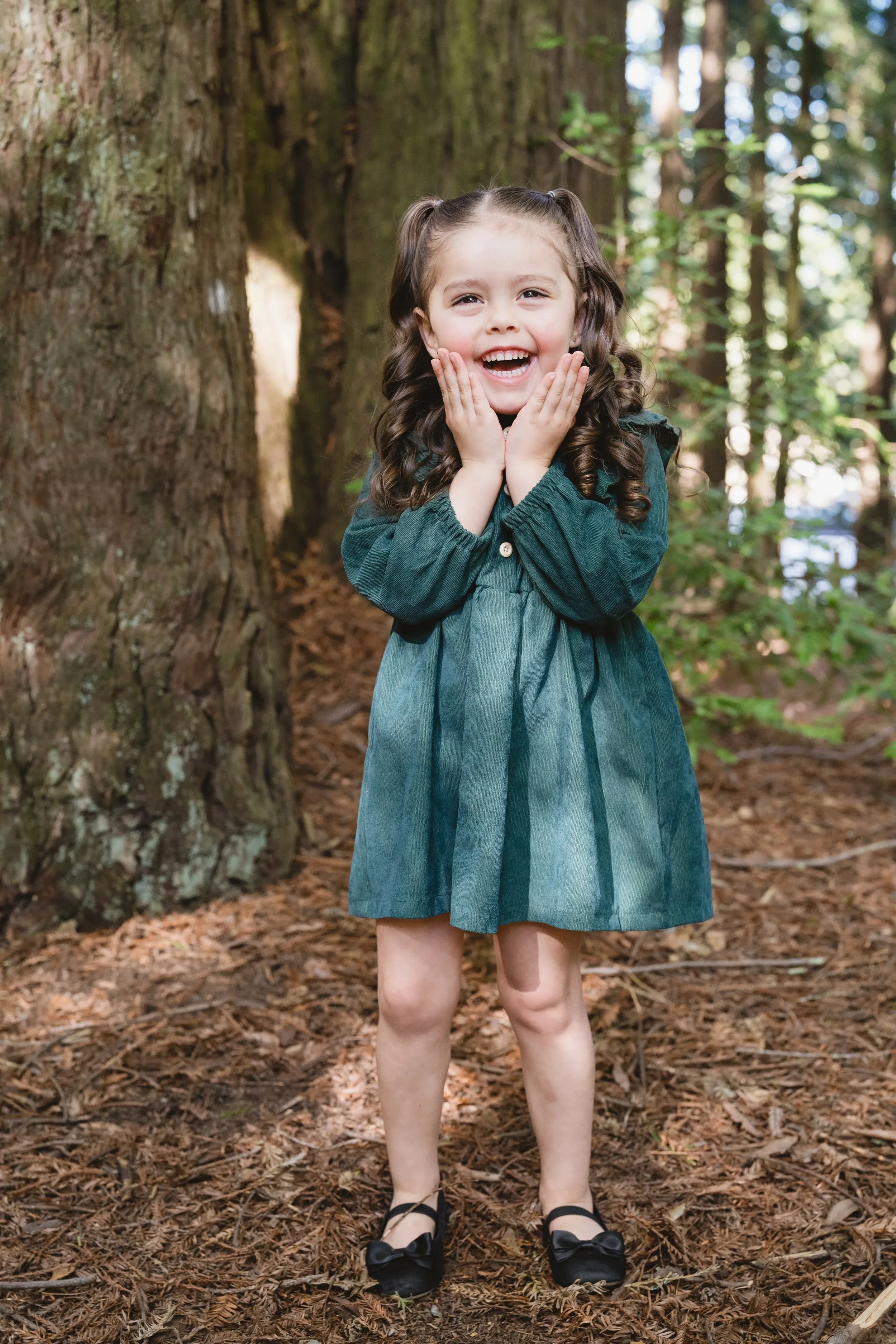 Full-length portrait of a young girl in a green corduroy dress laughing with both hands pressed to her cheeks on a redwood-needle forest floor