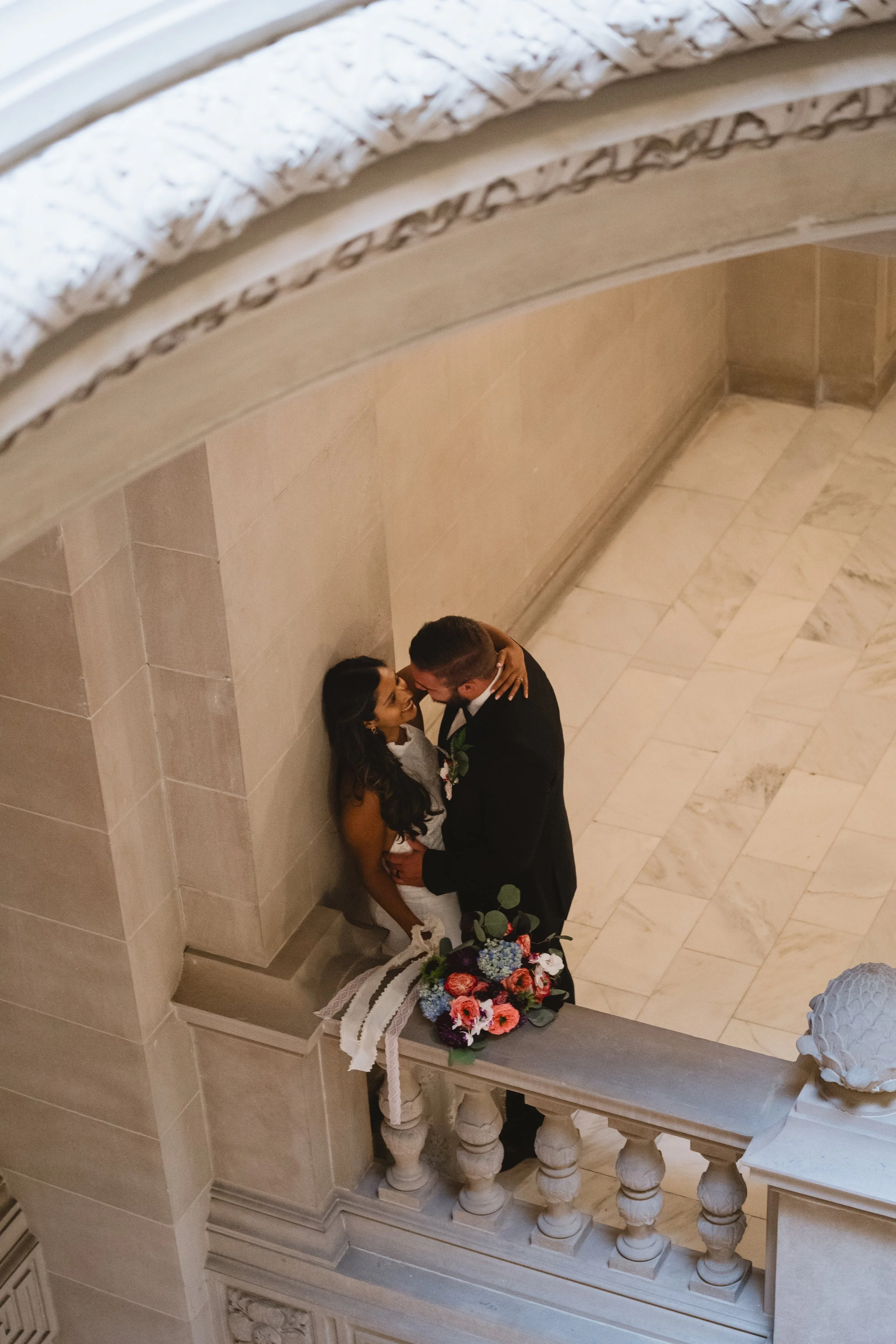 Aerial view of a couple embracing against a marble wall on an upper balcony landing inside City Hall.