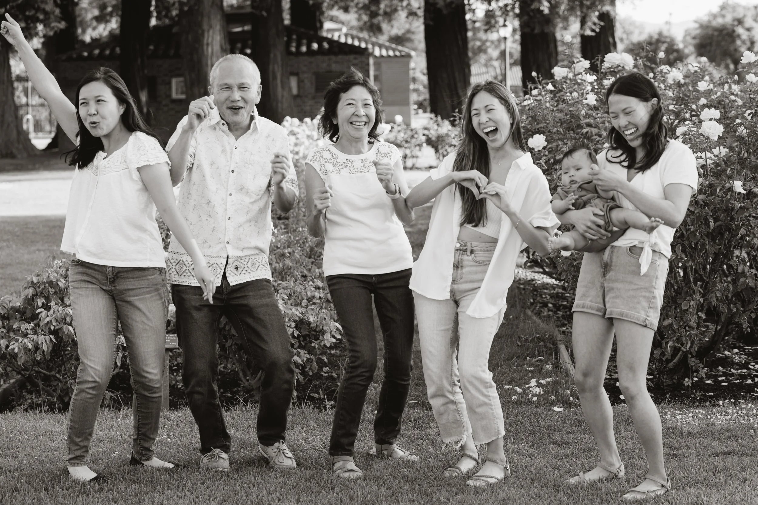 Group of five people, three women, one man, and a child, holding hands and smiling outdoors in a garden or park with trees and flowers in the background, in black and white.