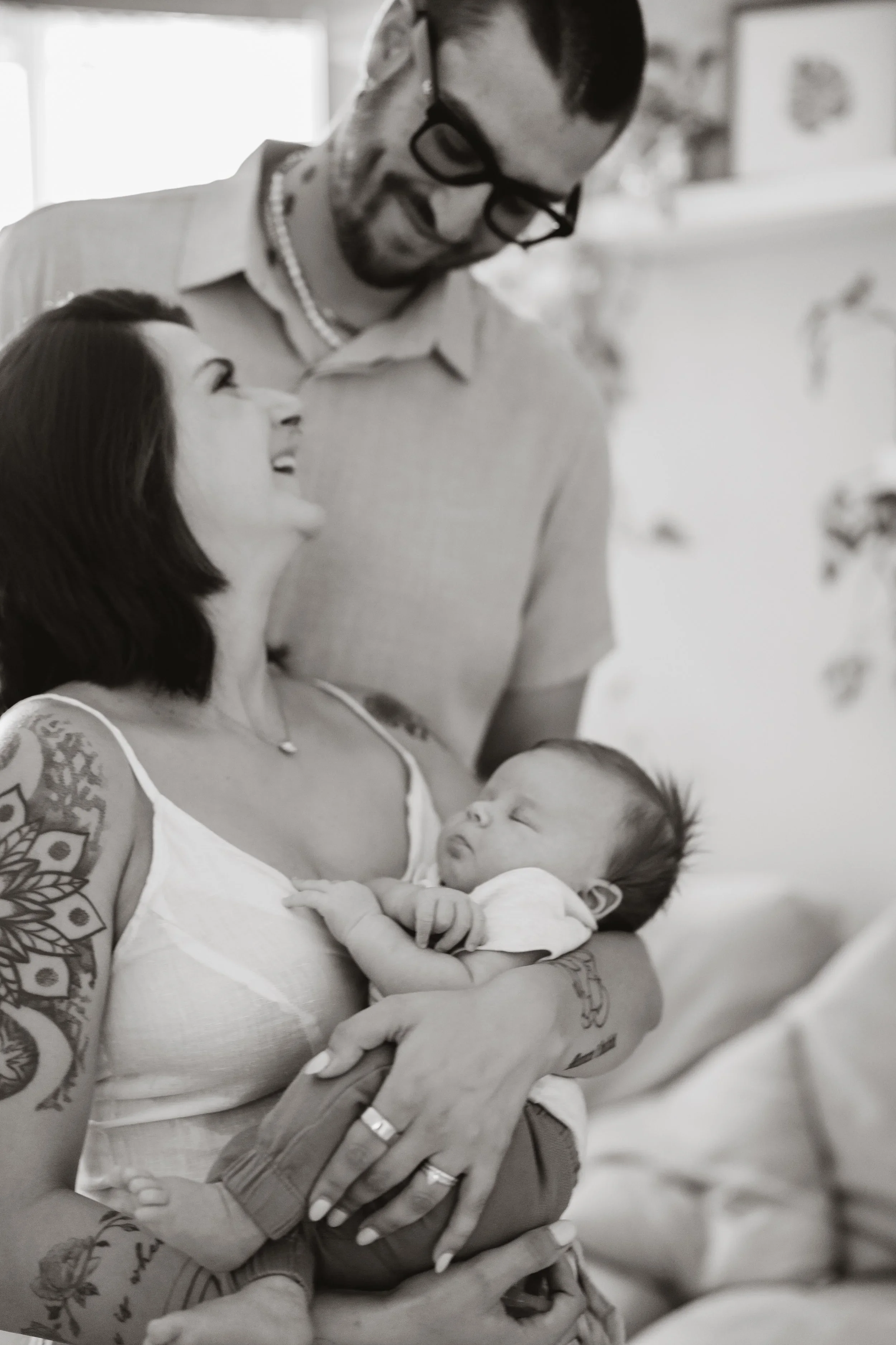 Black and white frame of a parent cradling a sleeping newborn and looking up at their partner, who leans in close from behind