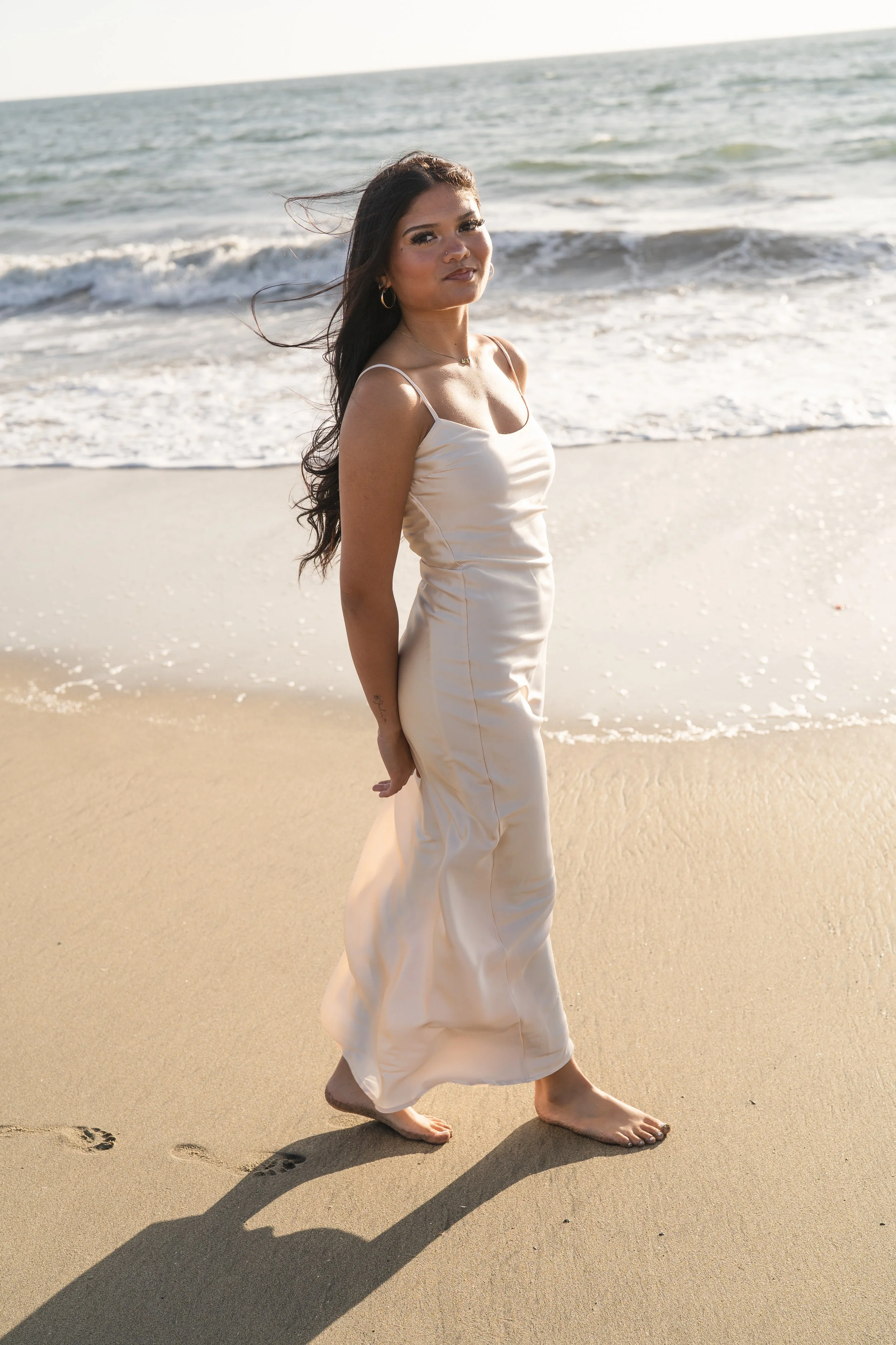 Senior girl walking barefoot along the shoreline at golden hour, glancing back at the camera in Half Moon Bay