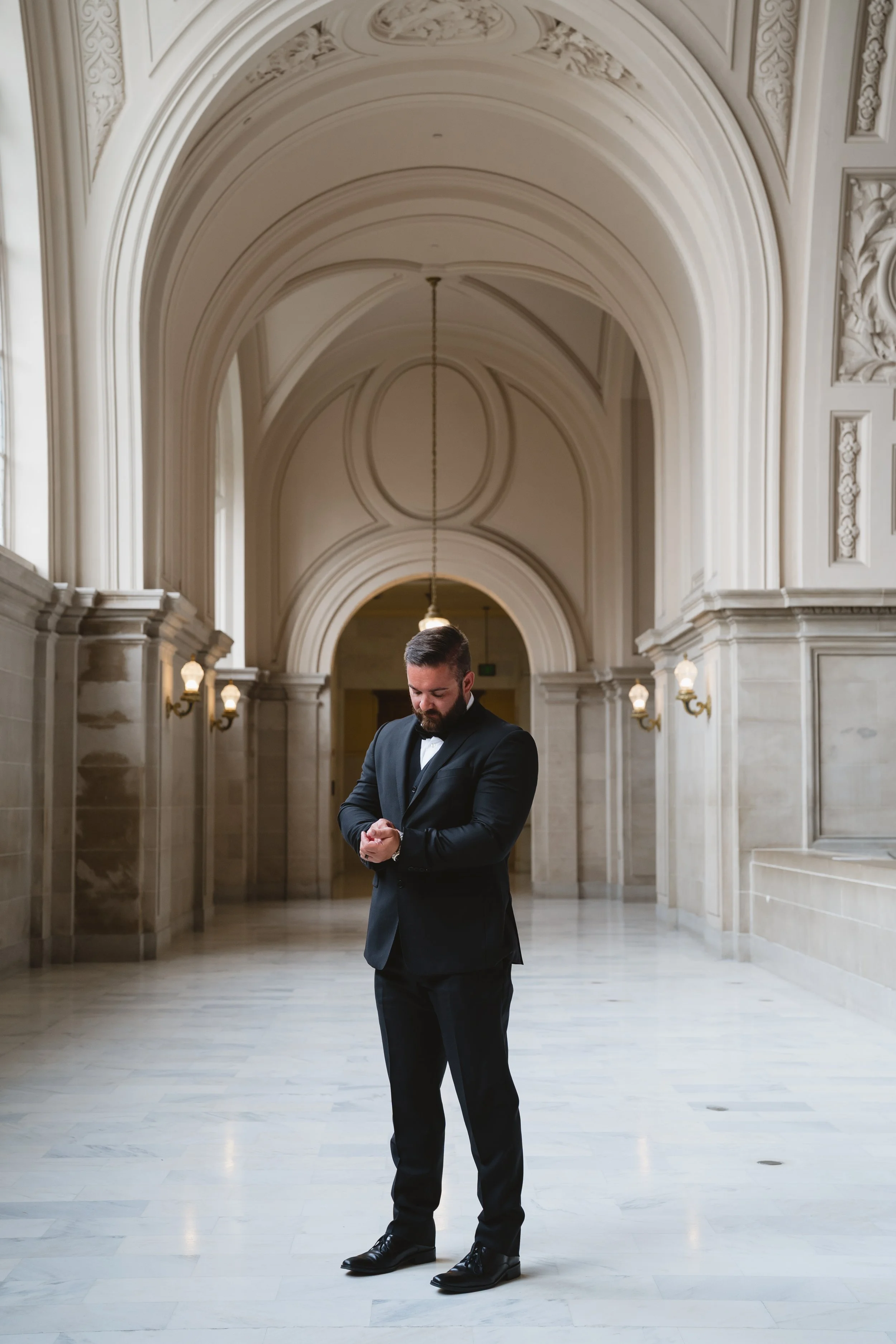 Groom standing alone in a vaulted marble corridor, adjusting his cufflink.