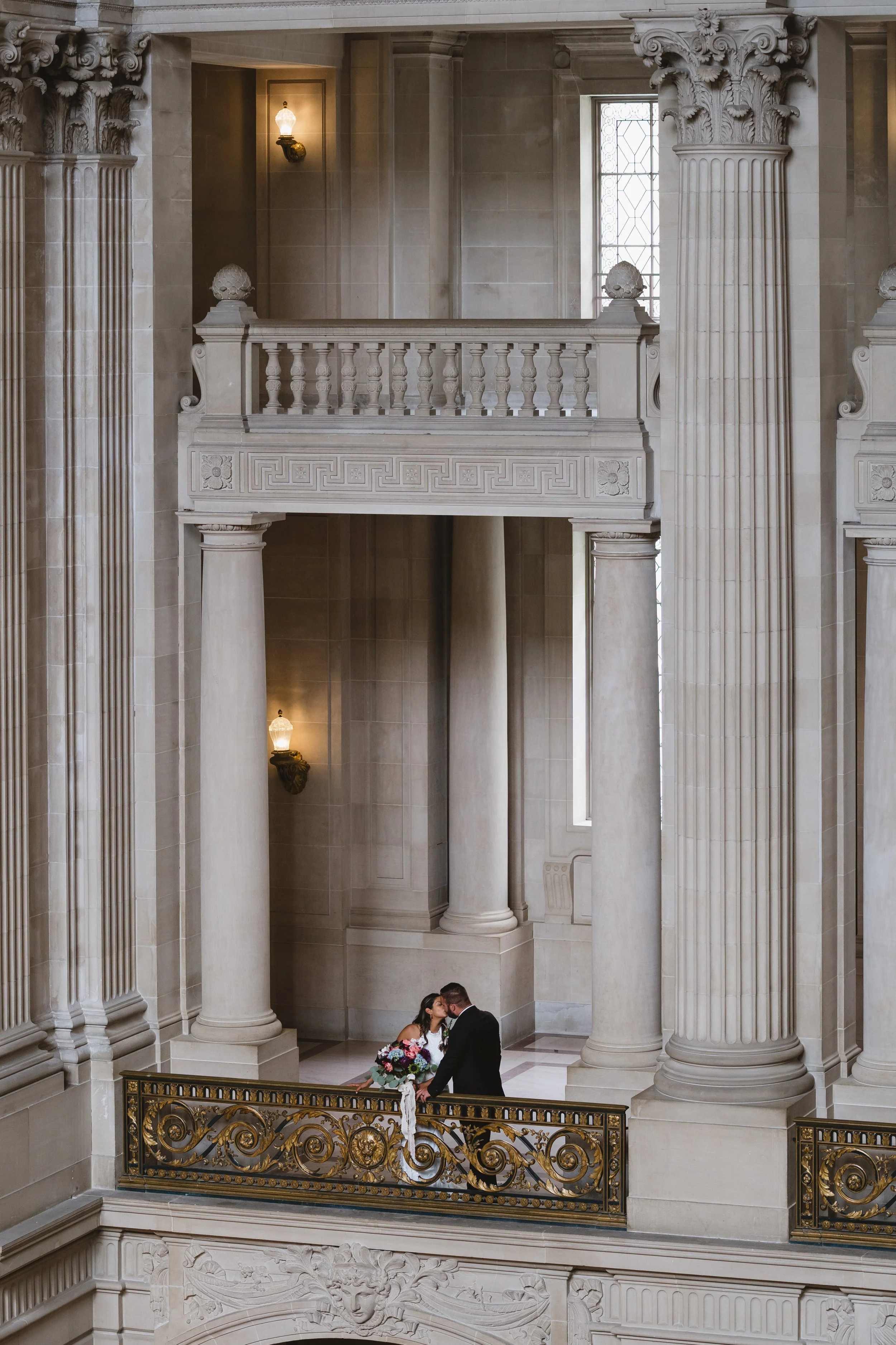 Couple kissing on a marble balcony inside San Francisco City Hall, seen from below through a row of columns.