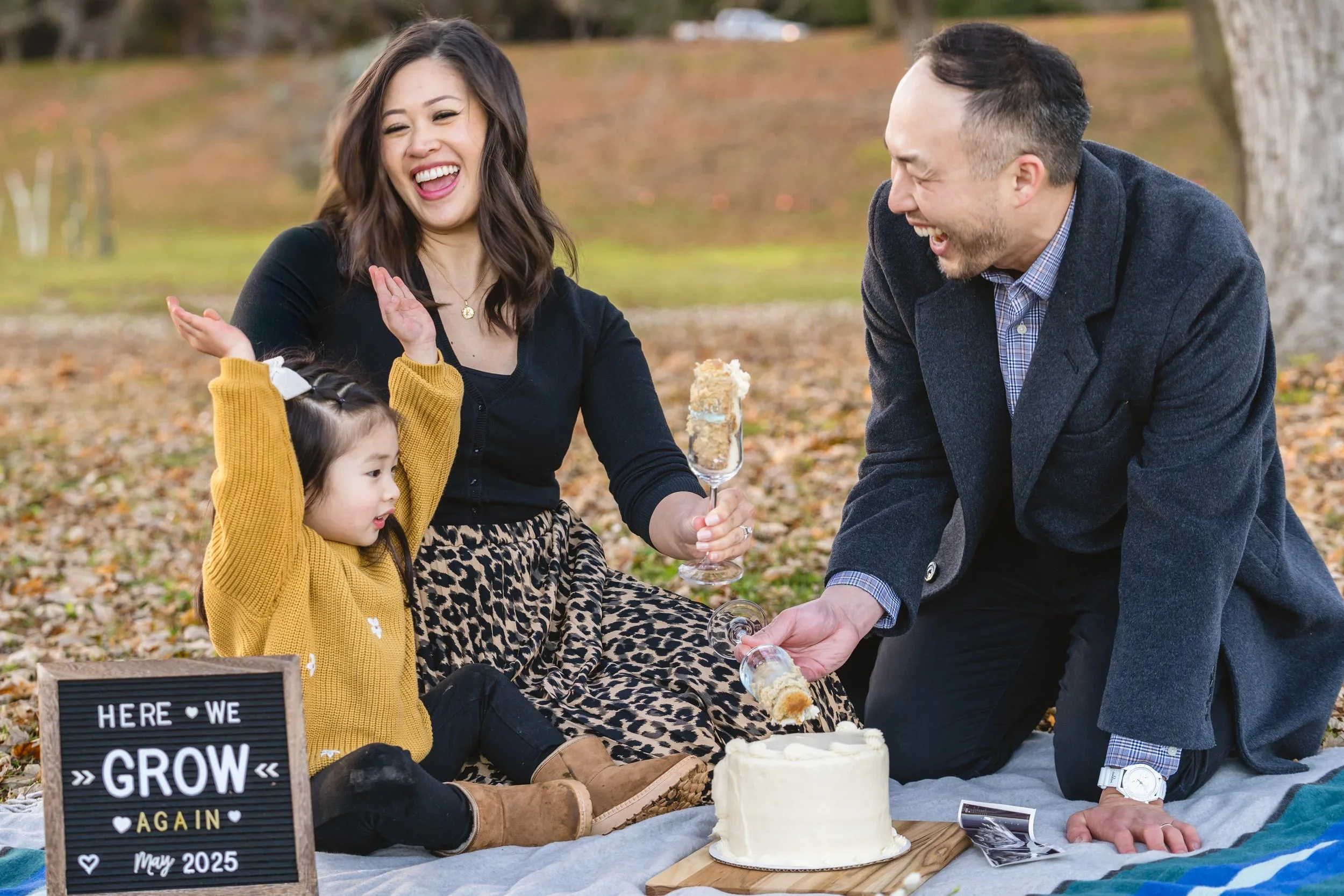 On a picnic blanket in fall leaves, a girl raises her arms while her parents laugh, holding glasses of cake beside a "Here We Grow Again" sign.