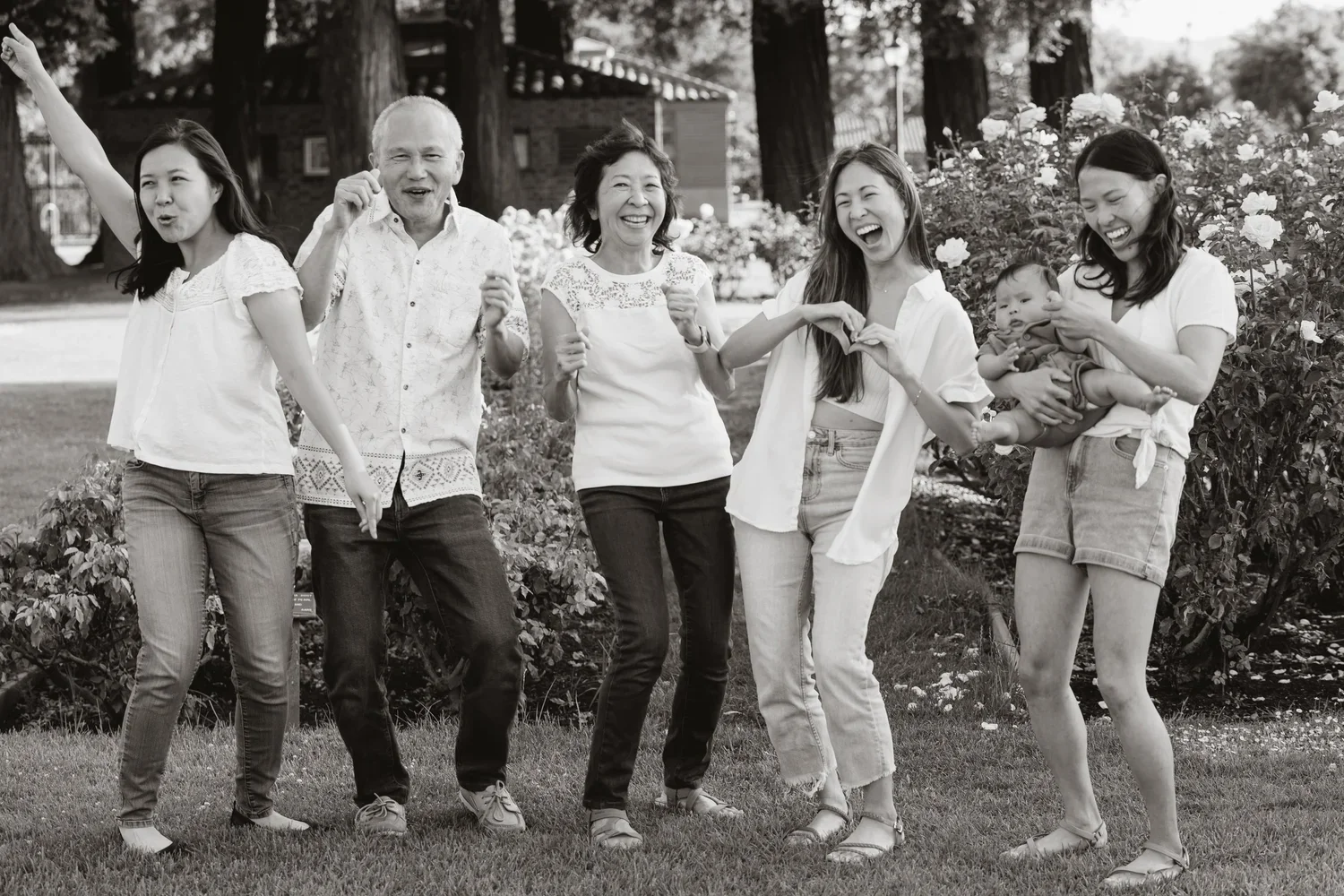 Three generations stand in a row dancing on the lawn in a black and white portrait, one daughter making a heart with her hands and another holding the baby on her hip.